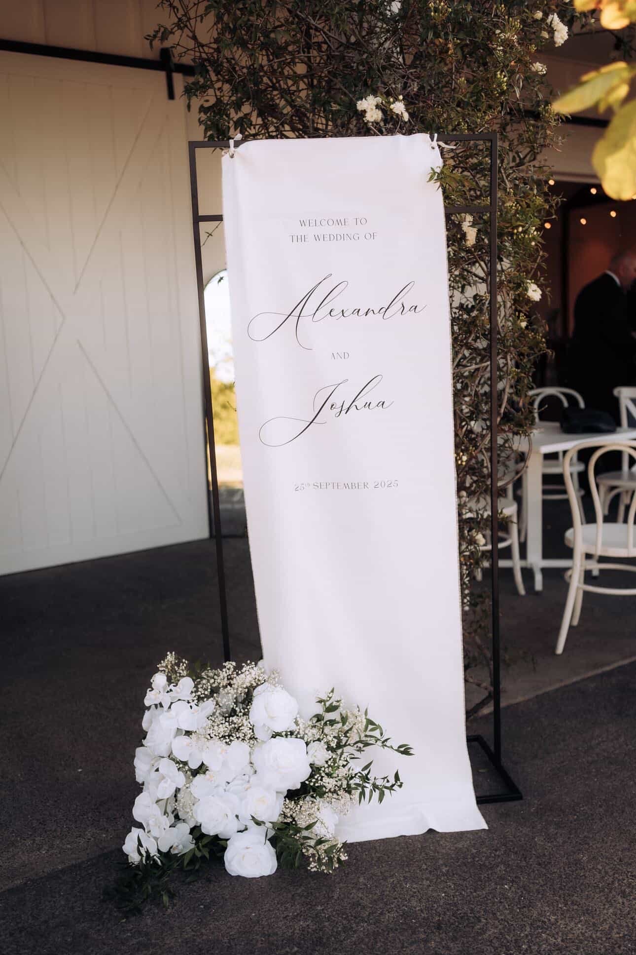 A wedding welcome sign with elegant black script text on a white cloth banner, decorated with white flowers and greenery, set outdoors near tables and chairs.
