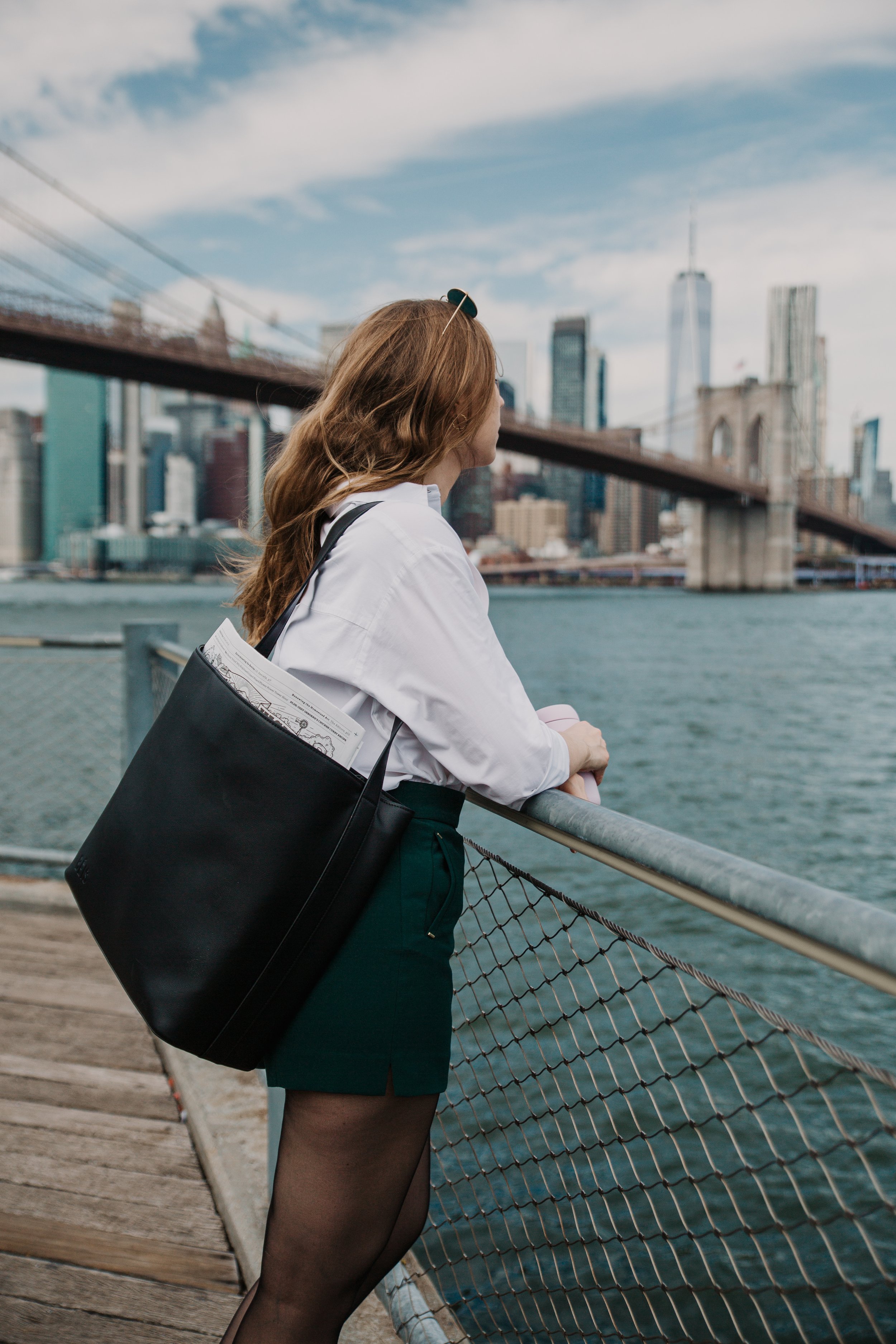 A woman with long wavy red hair standing on a wooden pier, leaning on a metal railing overlooking a body of water, with the Brooklyn Bridge and Manhattan skyline in the background. Lena Stalmasińska, Brand Strategy Consultant in Brooklyn, New York.
