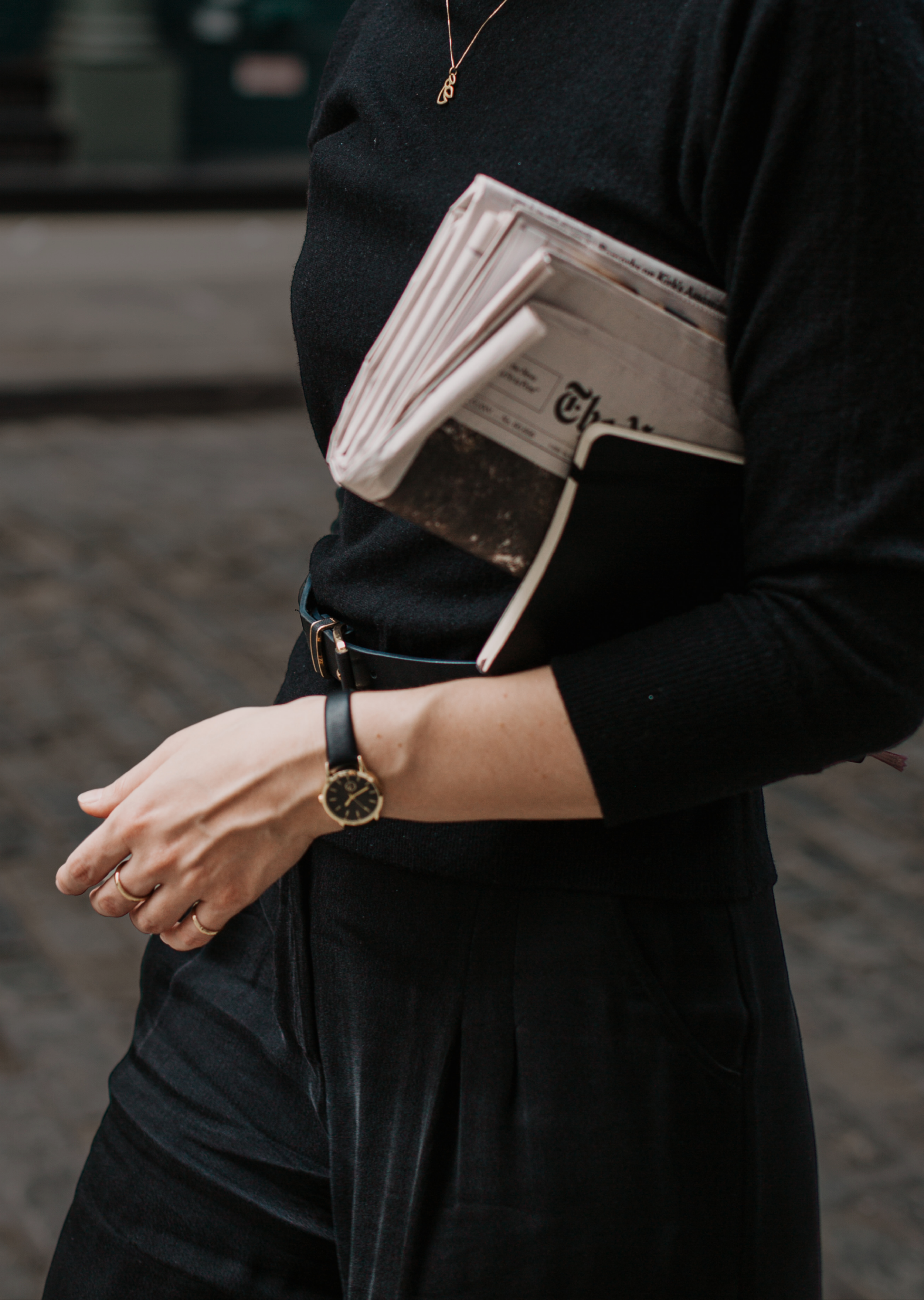 Person holding a stack of newspapers and a notebook, wearing a black long-sleeve top, gold watch, and black pants, standing outdoors.  Lena Stalmasińska, Brand Strategy Consultant in SOHO, New Your City.