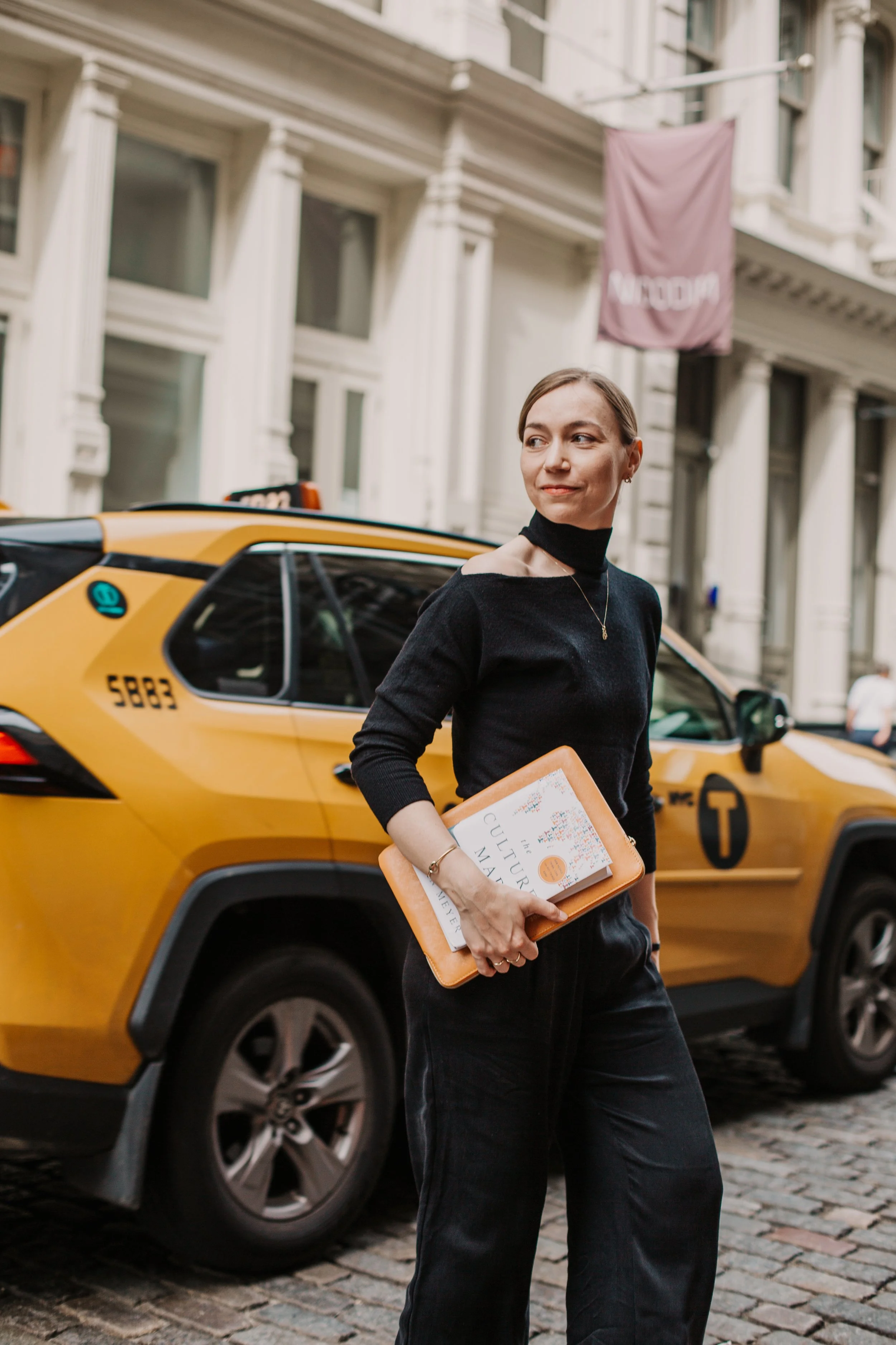 A woman with short hair holding a stack of books, walking on a cobblestone street next to a yellow taxi cab. She is wearing a black top and black pants, with a necklace and bracelet, and is looking sideways. In the background, there are old buildings with white facades and a purple banner hanging.