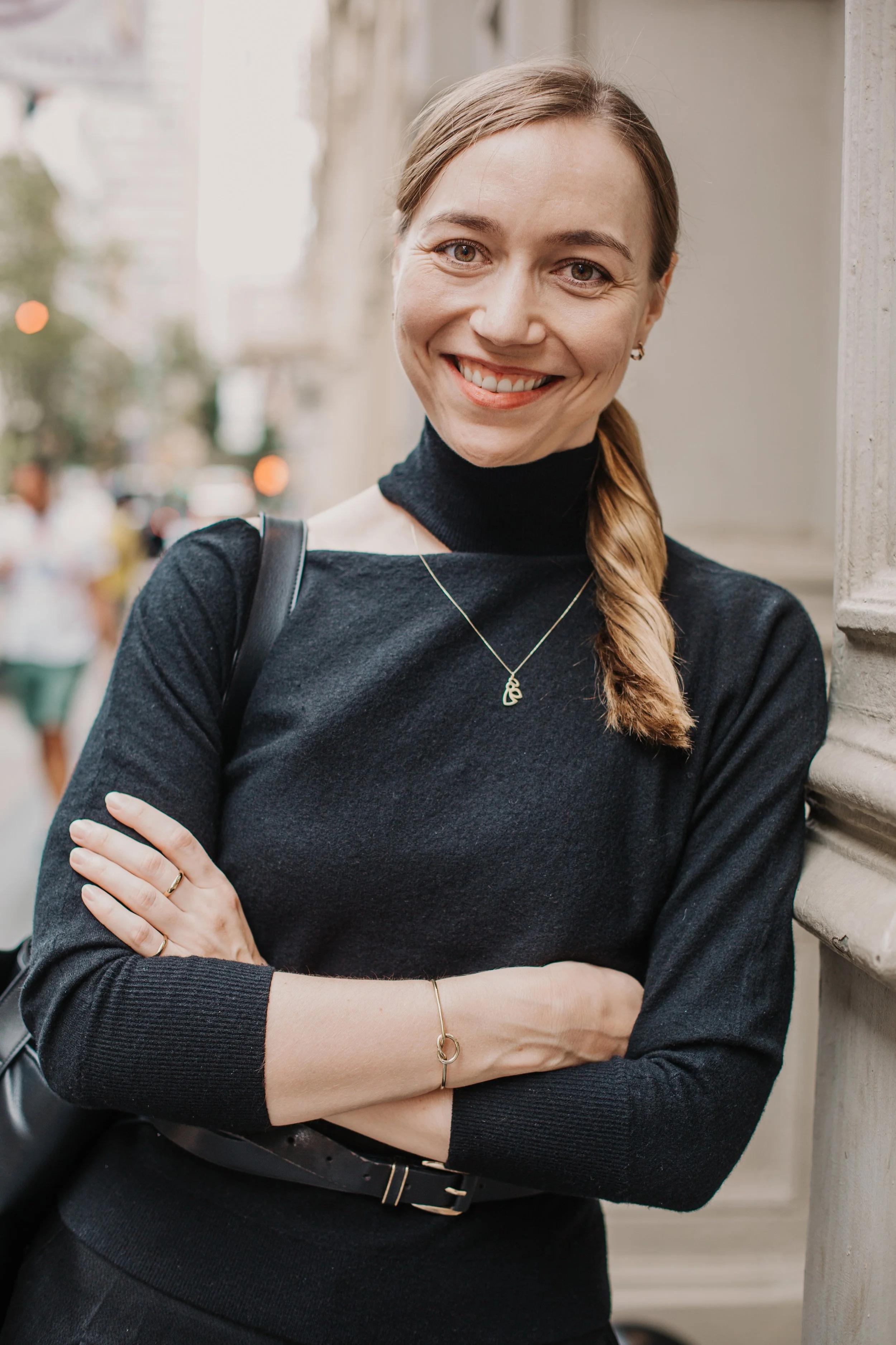 A woman with a braid, smiling, standing outdoors on a city street, wearing a black long-sleeve top and gold jewelry. Lena Stalmasińska, Brand Strategy Consultant in SOHO, New York City.