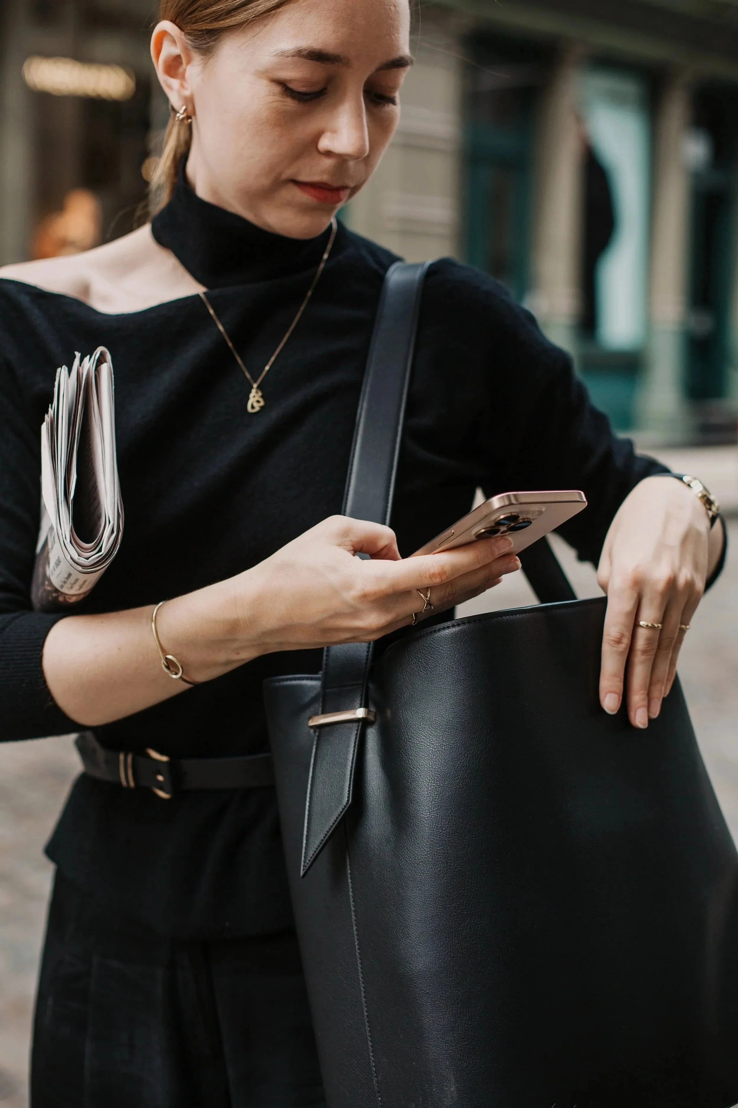 A woman with a black long-sleeve top, gold jewelry, and a black shoulder bag, holding a smartphone and a rolled-up magazine in an urban setting. Lena Stalmasińska, Brand Strategy Consultant in SOHO, New York City.