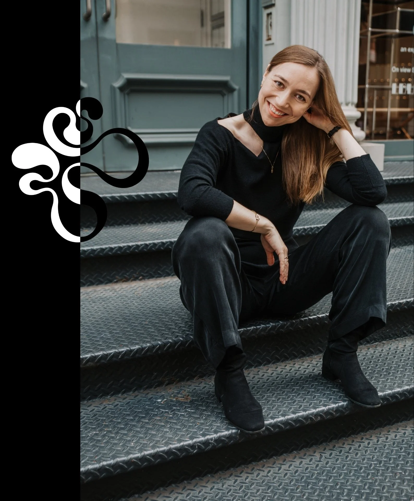 A woman with long brown hair, wearing a black sweater and black pants, sitting on metal stairs outside a building, smiling and leaning her head on her hand. Lena Stalmasińska, Brand Strategy Consultant in SOHO, New York City.