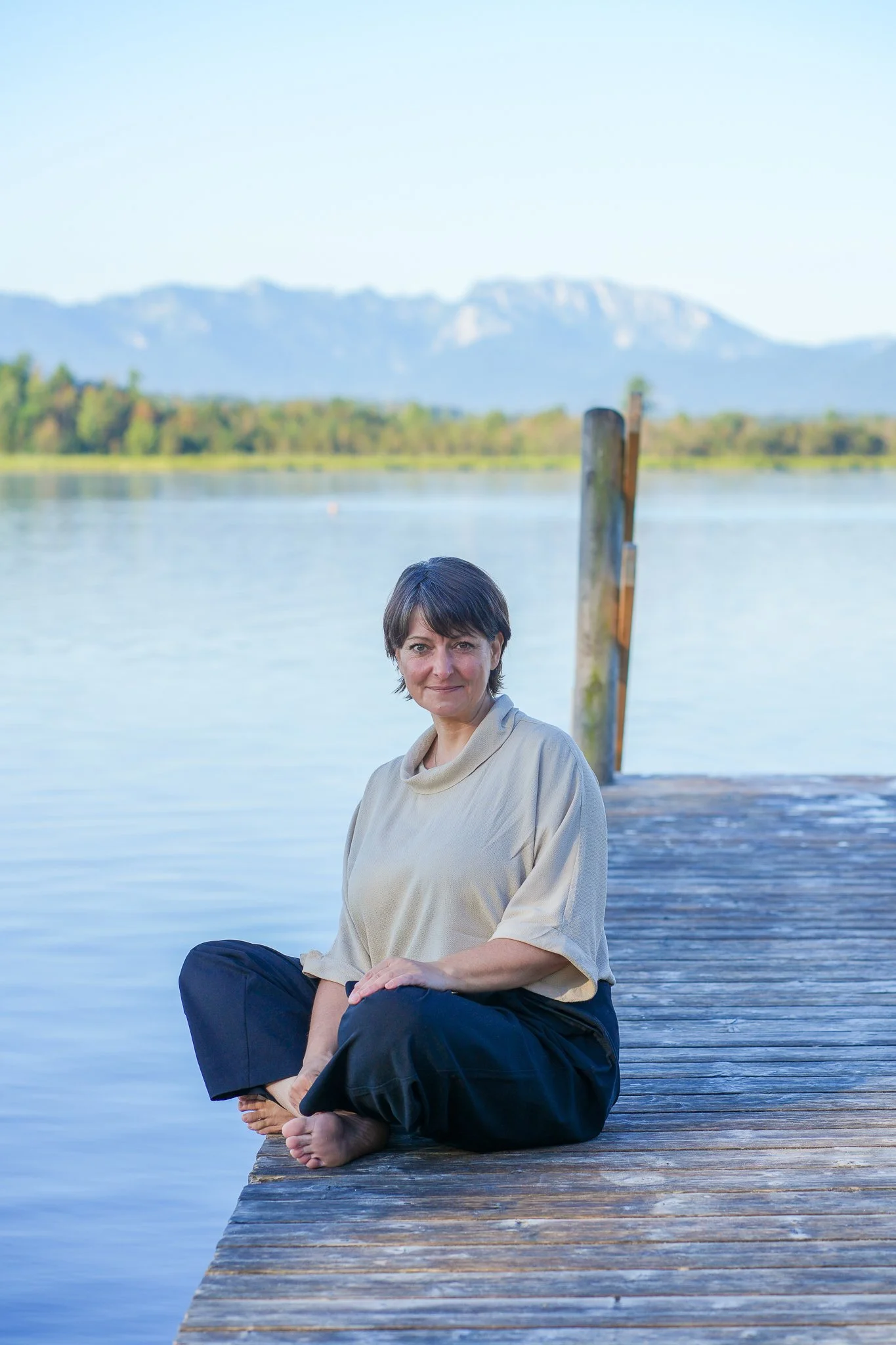 Bernadette sitzt entspannt an einem Steg am Wasser mit Bergen im Hintergrund.