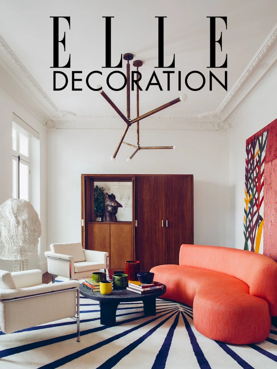 Salon intérieur moderne avec canapés blancs et orange, tapis rayé noir et blanc, table basse noire avec des livres et des vaisselles colorées, armoire en bois, tableau abstrait coloré, et un chandelier mural moderne.