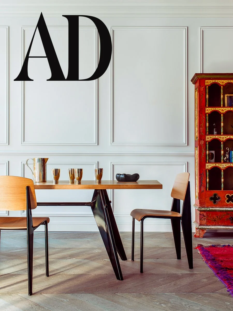 Une salle à manger élégante avec une table en bois clair, deux chaises en bois, une étagère rouge avec des accents dorés, et des objets décoratifs. La pièce a un mur blanc avec des moulures.