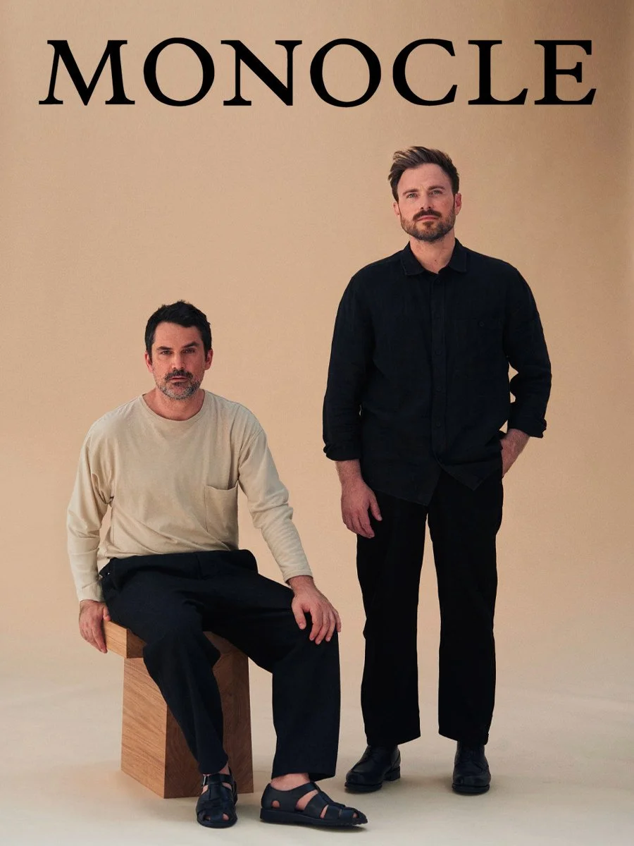 Deux hommes posent devant un fond neutre, l'un assis sur un cube en bois et l'autre debout à côté. Le titre 'MONOCLE' apparaît en haut de l'image.