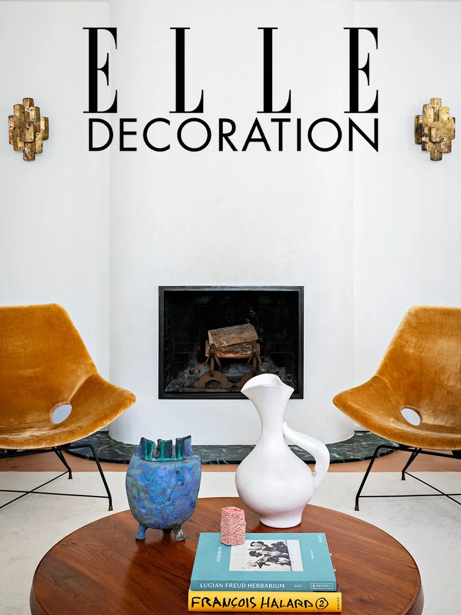 Salle de réception élégante avec une cheminée, deux fauteuils en velours orange, une table basse en bois avec plusieurs objets décoratifs, dont un vase blanc, un vase bleu, un livre sur Lucian Freud et un livre de François Halard, et deux sculptures 