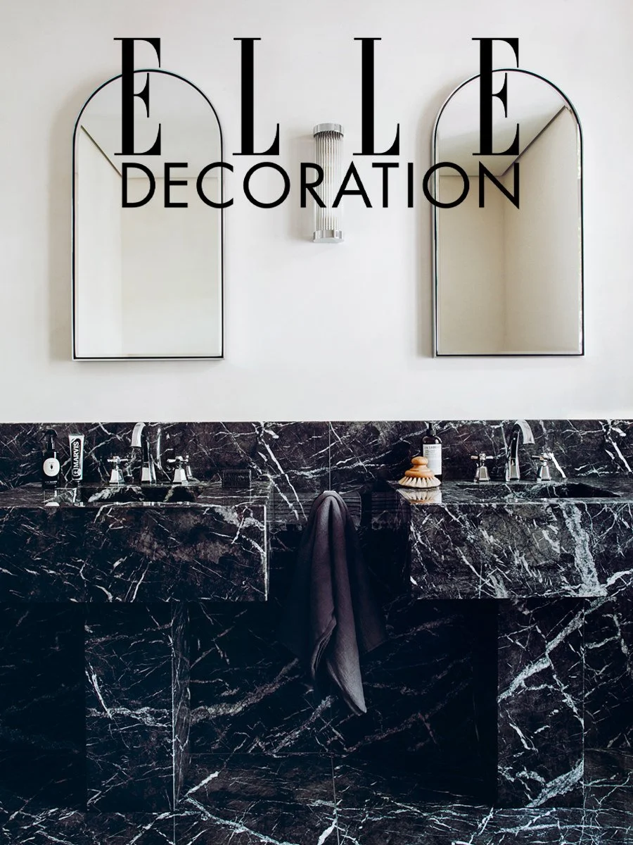Salle de bain avec double lavabo en marbre noir, deux miroirs en forme d'arc, et un mur blanc avec une décoration ELLE.