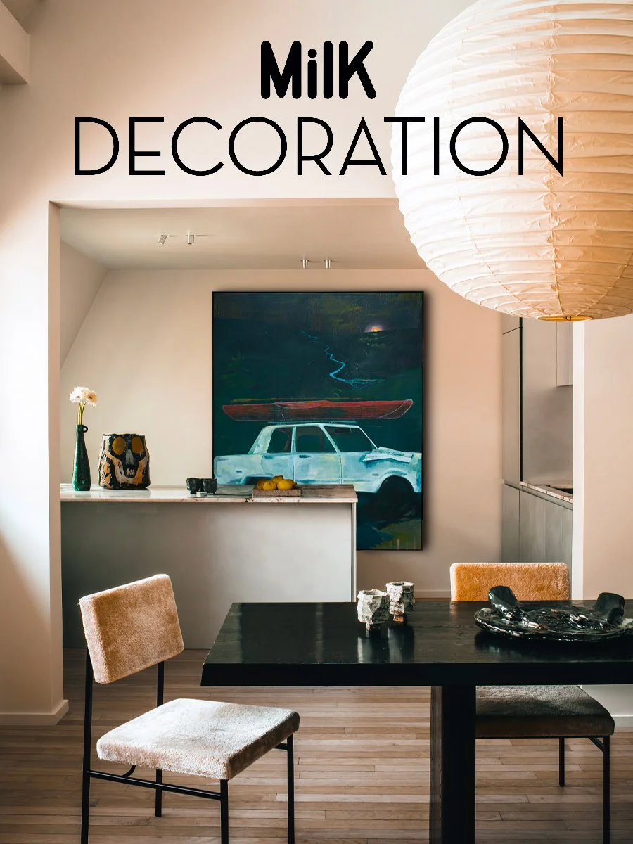 Une salle à manger moderne avec une table en bois noir, des chaises en tissu beige, une œuvre d'art représentant une voiture blanche avec un kayak rouge sur le toit, et une grande lampe ronde en papier blanc suspendue au plafond.