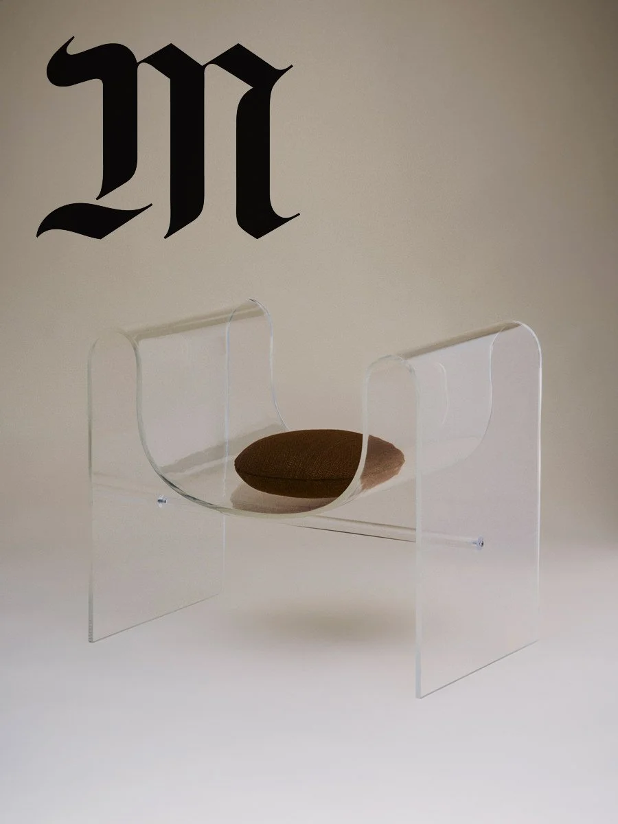 Une chaise transparente avec un coussin marron dessus, placée contre un mur blanc avec une grande lettre noire 'M' au-dessus.