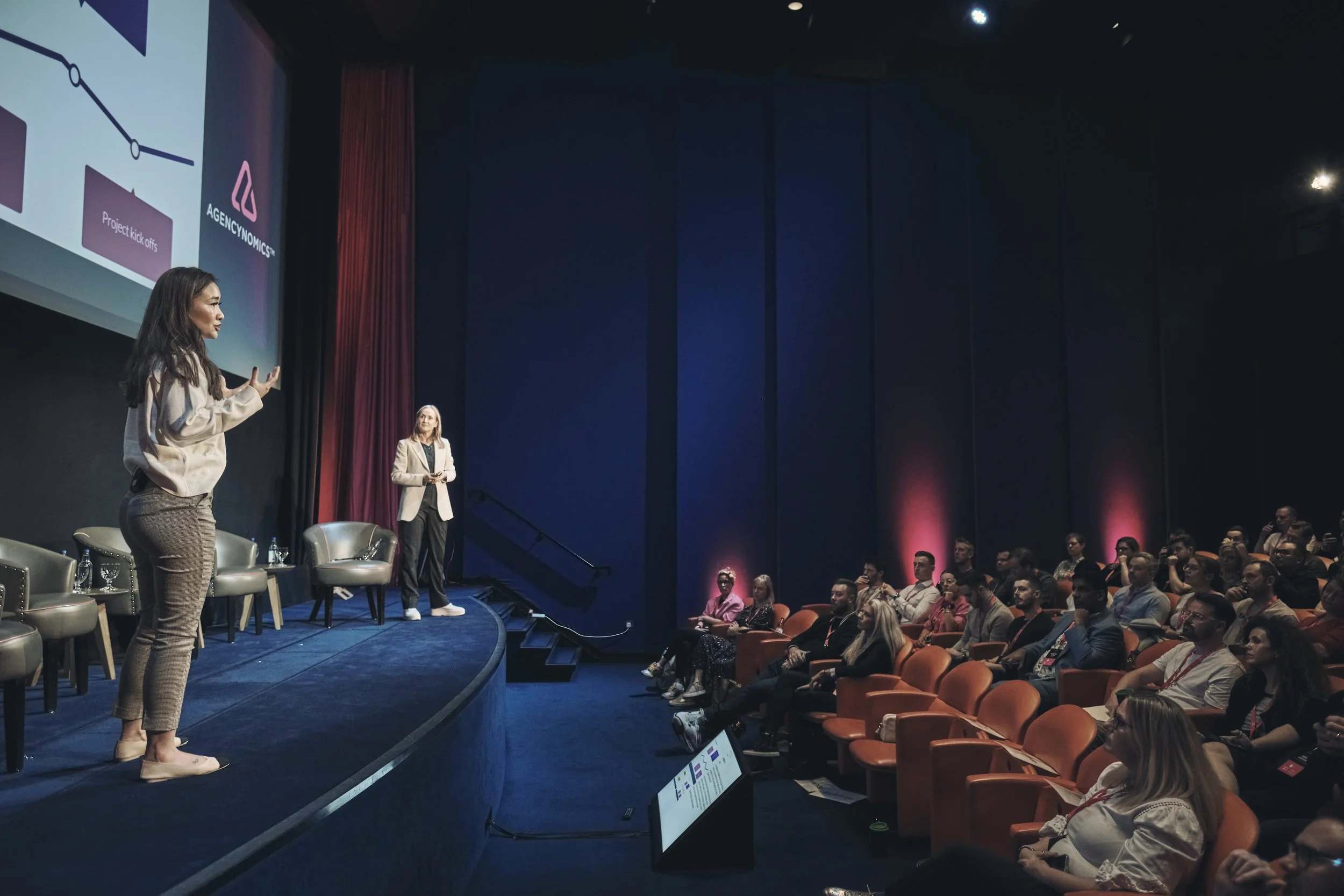 A woman presents on stage in front of a seated audience during a conference or seminar, with a large screen displaying a PowerPoint presentation behind her.