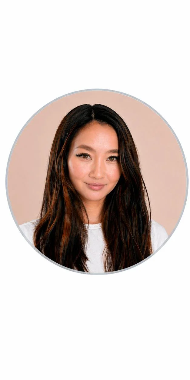 A woman with long dark hair and subtle makeup, wearing a white top, looking at the camera with a slight smile, against a pale background.
