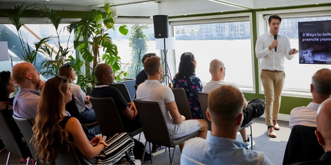 A man giving a presentation to a group of people in a modern, bright room with large windows and plants.