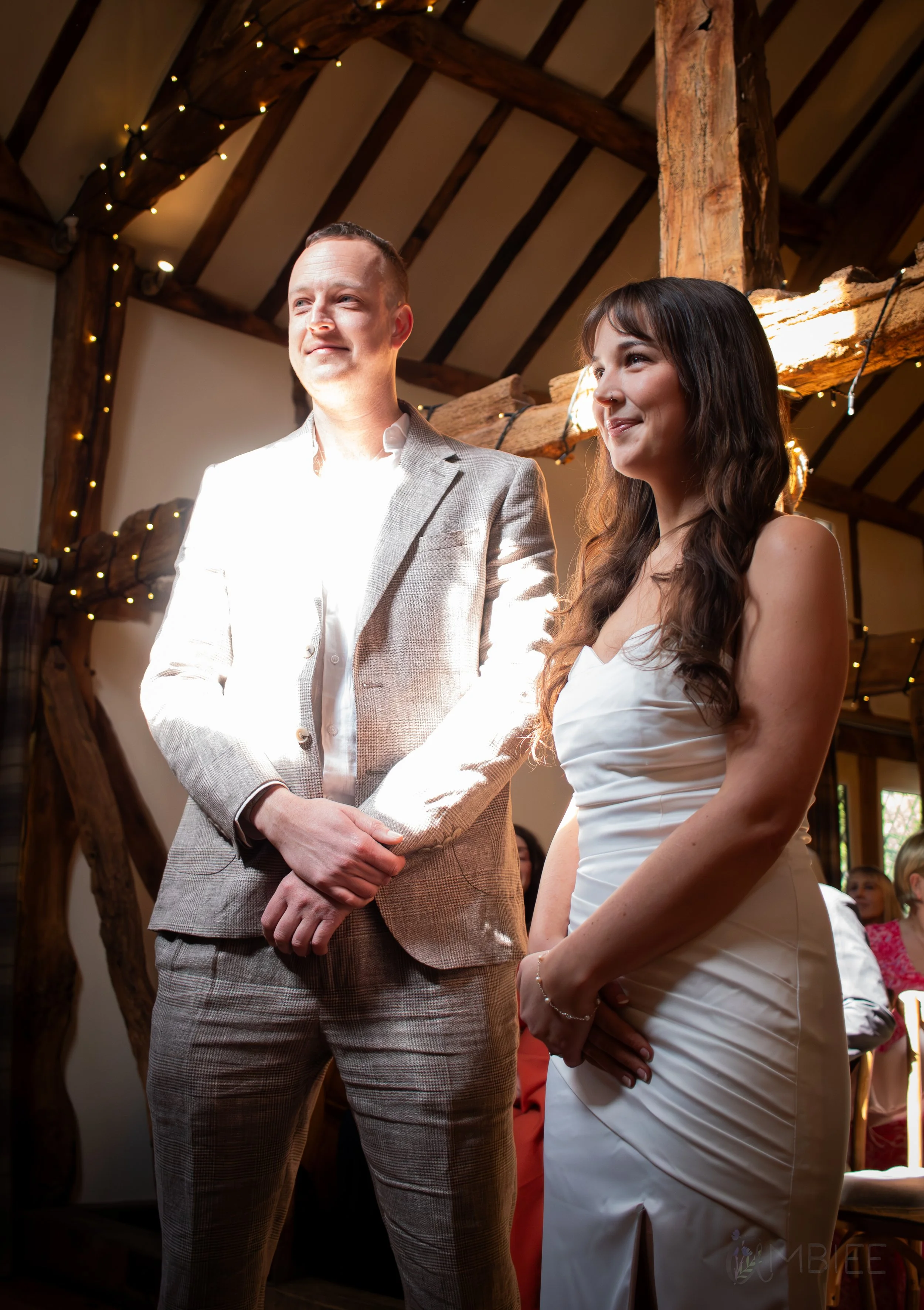 A couple standing during a wedding ceremony indoors, with the man in a light-colored plaid suit and the woman in a white wedding dress, with warm lighting and wood beams overhead.