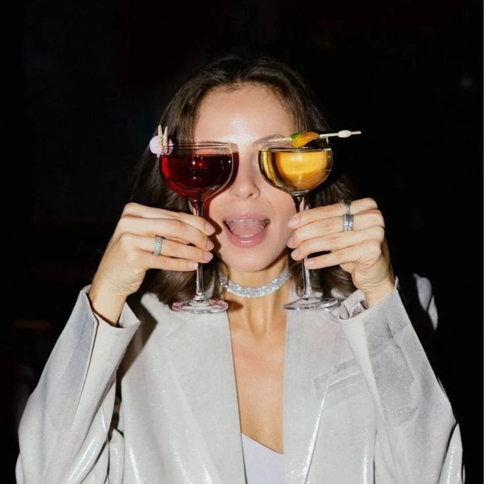 Woman holding two cocktails up to her eyes, one red and one yellow, with garnishes, in a dark setting