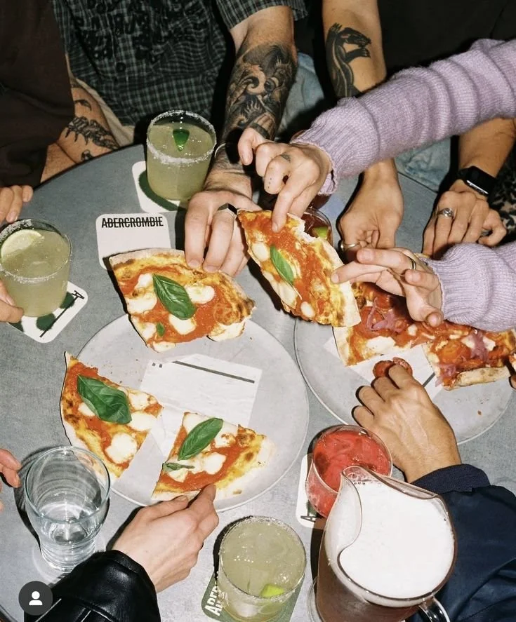 Group of people at a table sharing pizza and drinks. Four pizza slices on plates, some with basil leaves. Several glasses with beverages, some garnished with lime. Hands reaching for pizza slices.