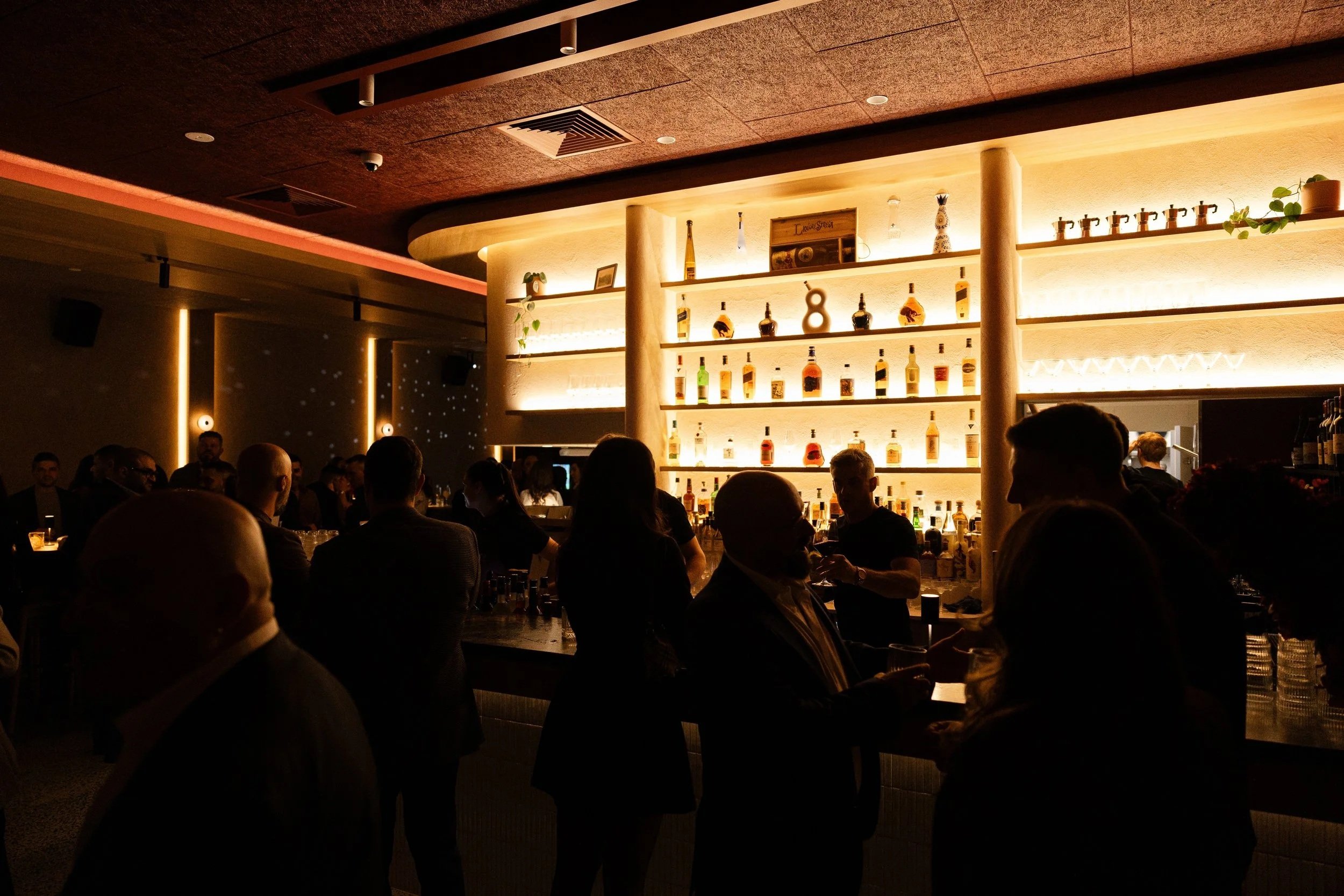 A busy bar scene with patrons sitting and standing, illuminated shelves with bottles of alcohol, and a warm ambient lighting.
