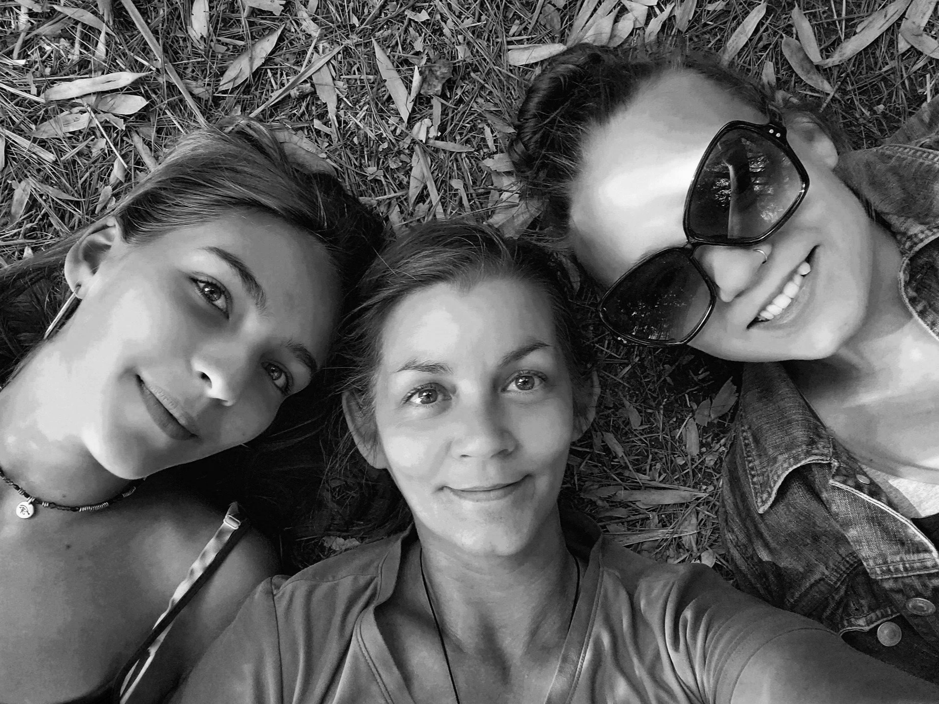 Three women lying on the ground outdoors, with leaves and grass around them, taking a selfie.