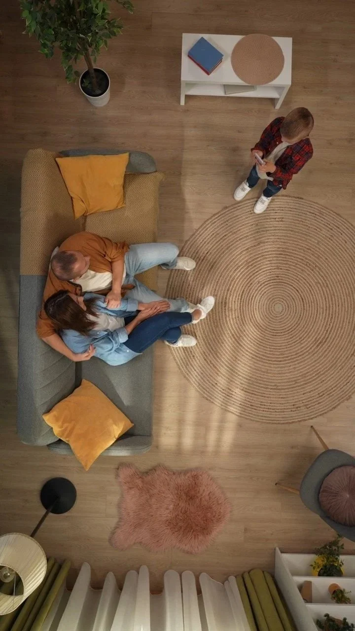 A top-down view of a living room with two people sitting on a sofa and a child standing nearby. The room has a wooden floor, a round beige rug, potted plant, bookshelf, and a side table with books.