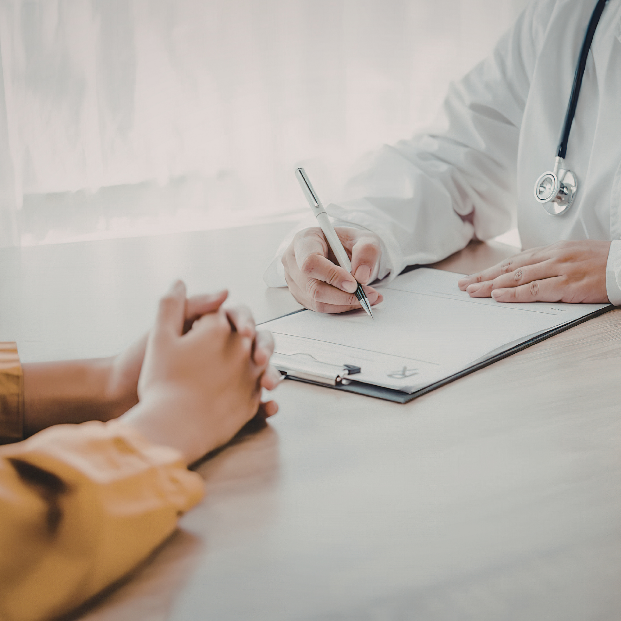 A healthcare professional in a white coat and stethoscope writing on a clipboard while sitting at a desk, with a patient's hand visible in the foreground.