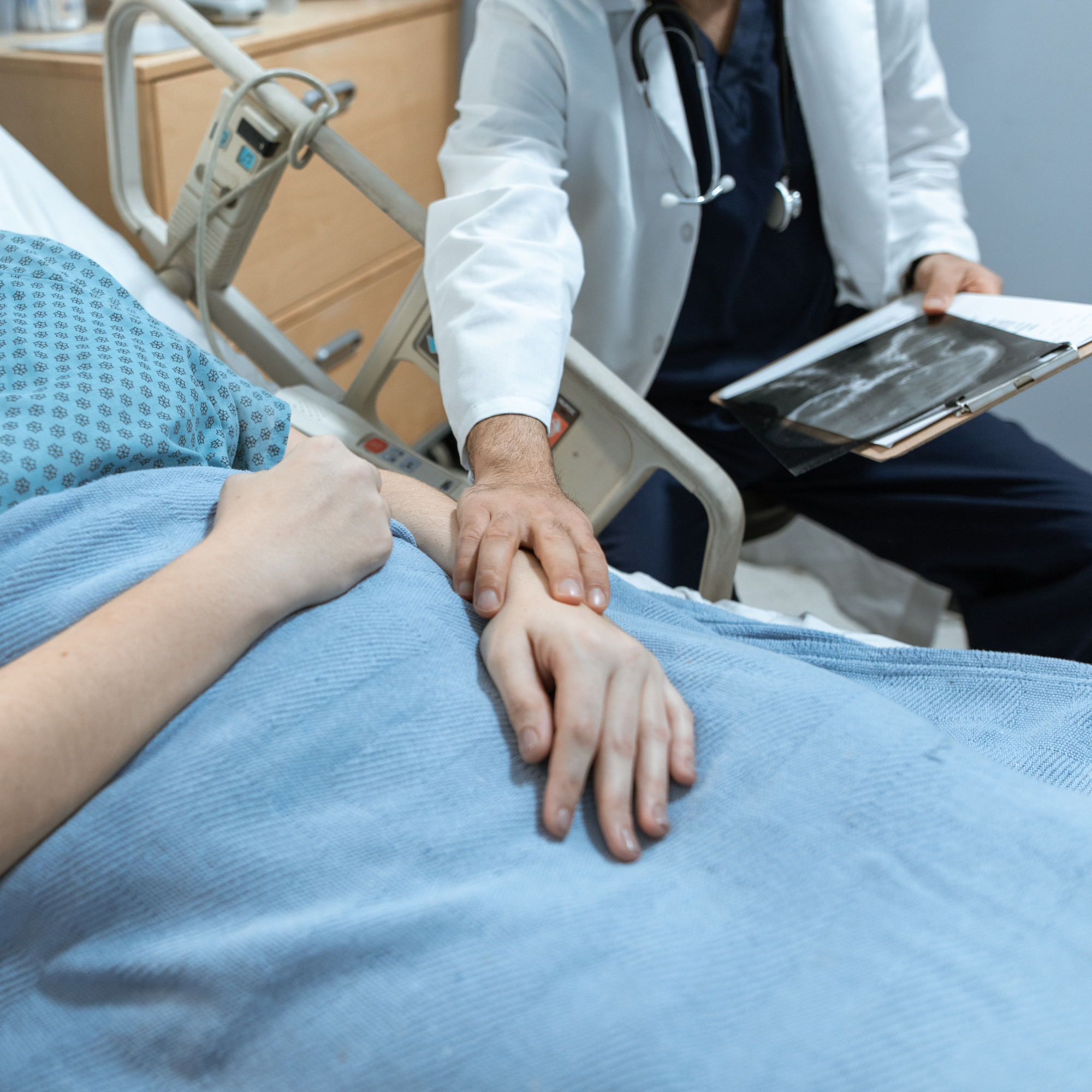 A doctor taking the patient's pulse with a stethoscope and holding an X-ray scan, patient lying on hospital bed with blue patterned sheet.