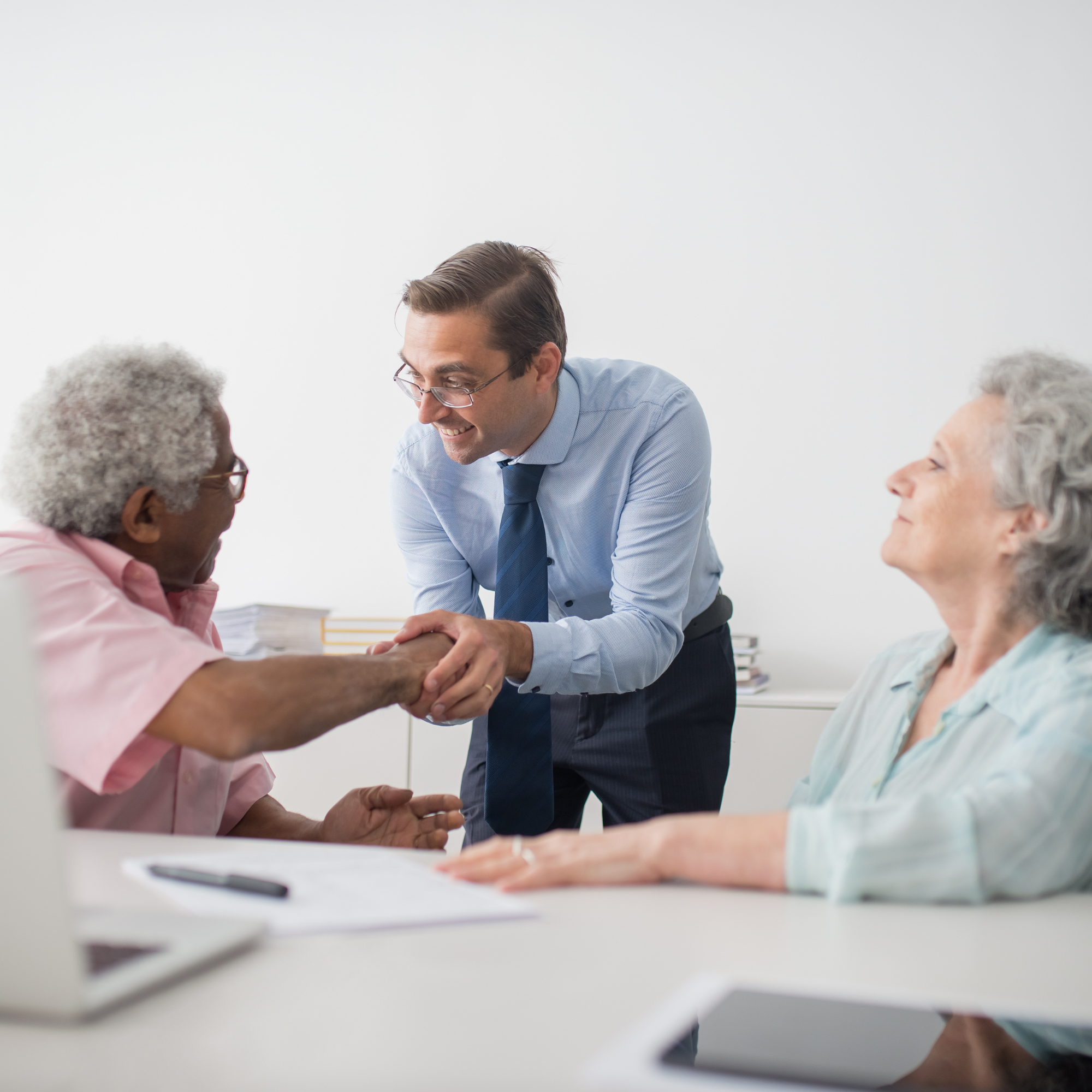 A young man with glasses and a blue shirt leaning forward and smiling, shaking hands with an older woman with gray hair, also smiling, in a room with white walls.