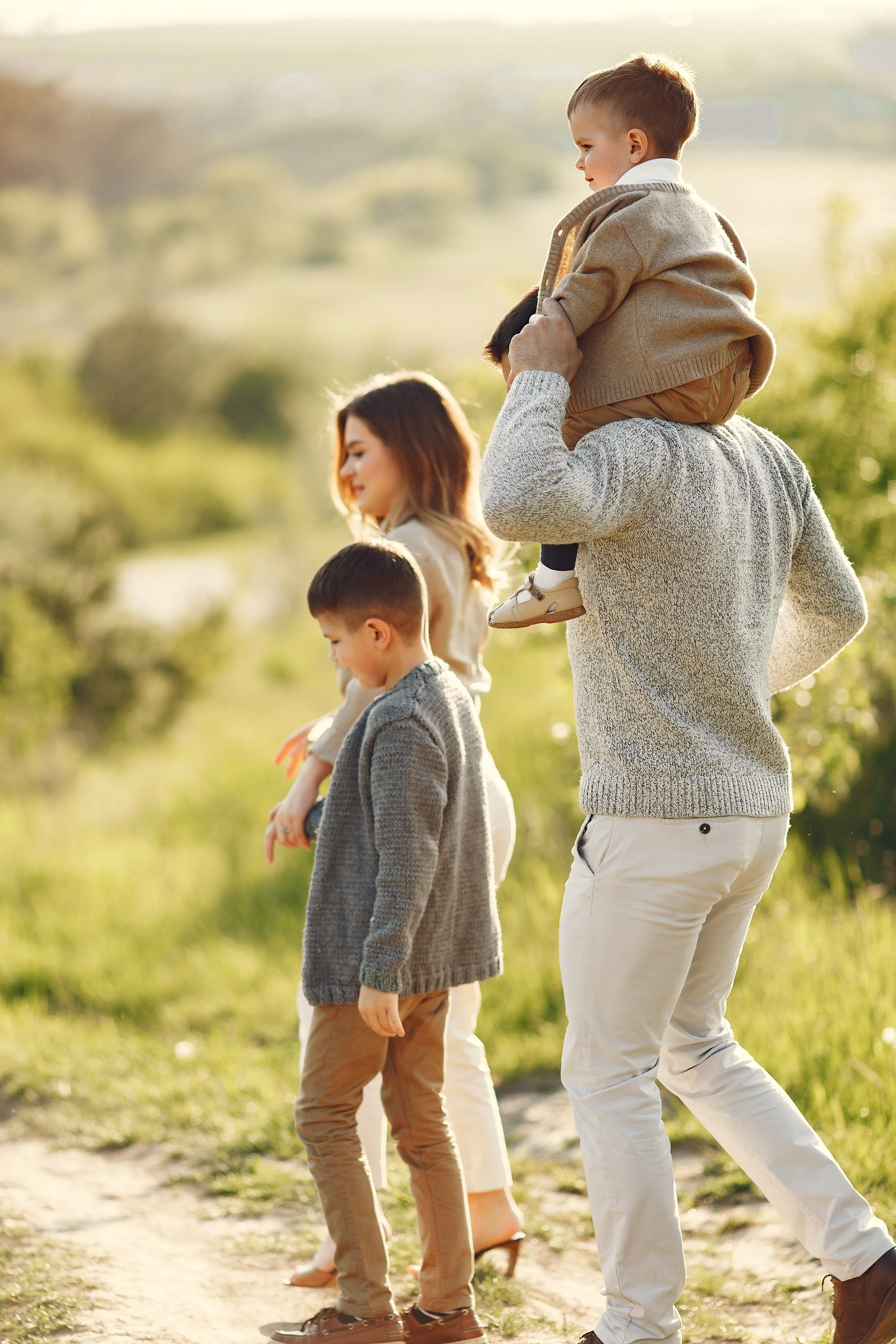A family walking outdoors in a sunlit green field, with a man carrying a young boy on his shoulders, and two other children walking alongside.