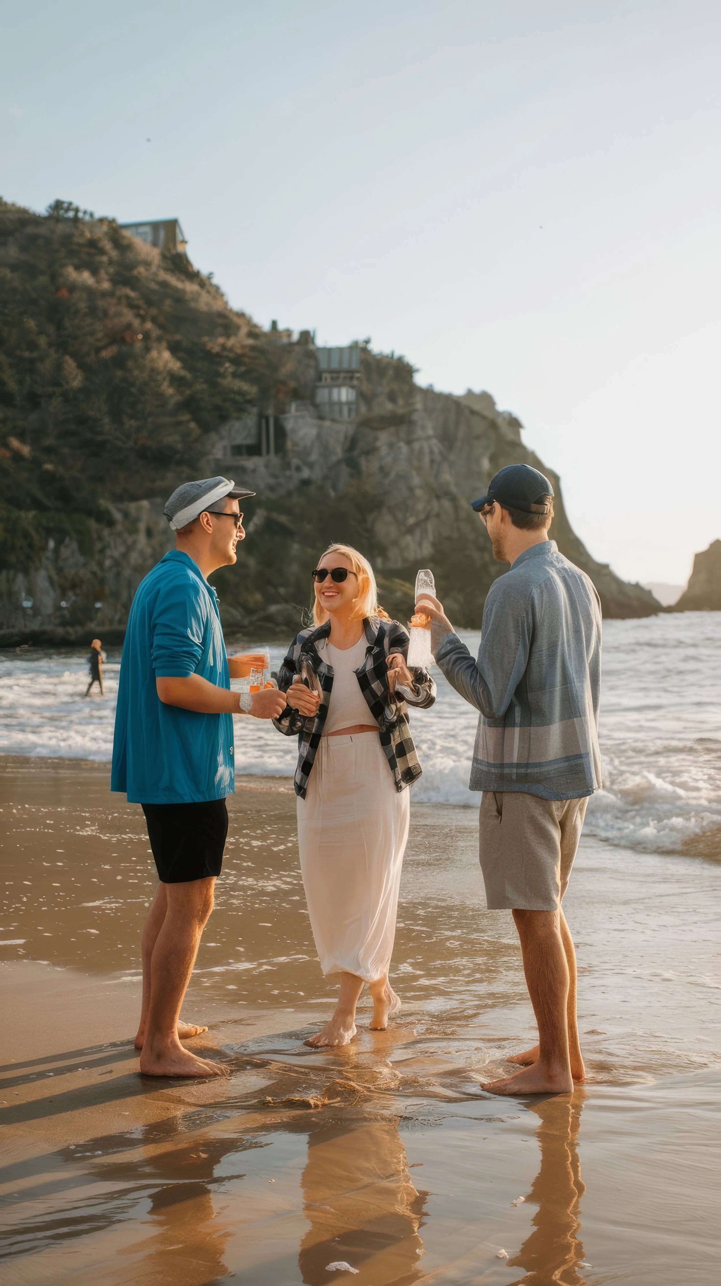 Three friends, two men and one woman, standing on the beach near the water, enjoying drinks and smiling at each other, with the ocean and cliffs in the background during sunset.