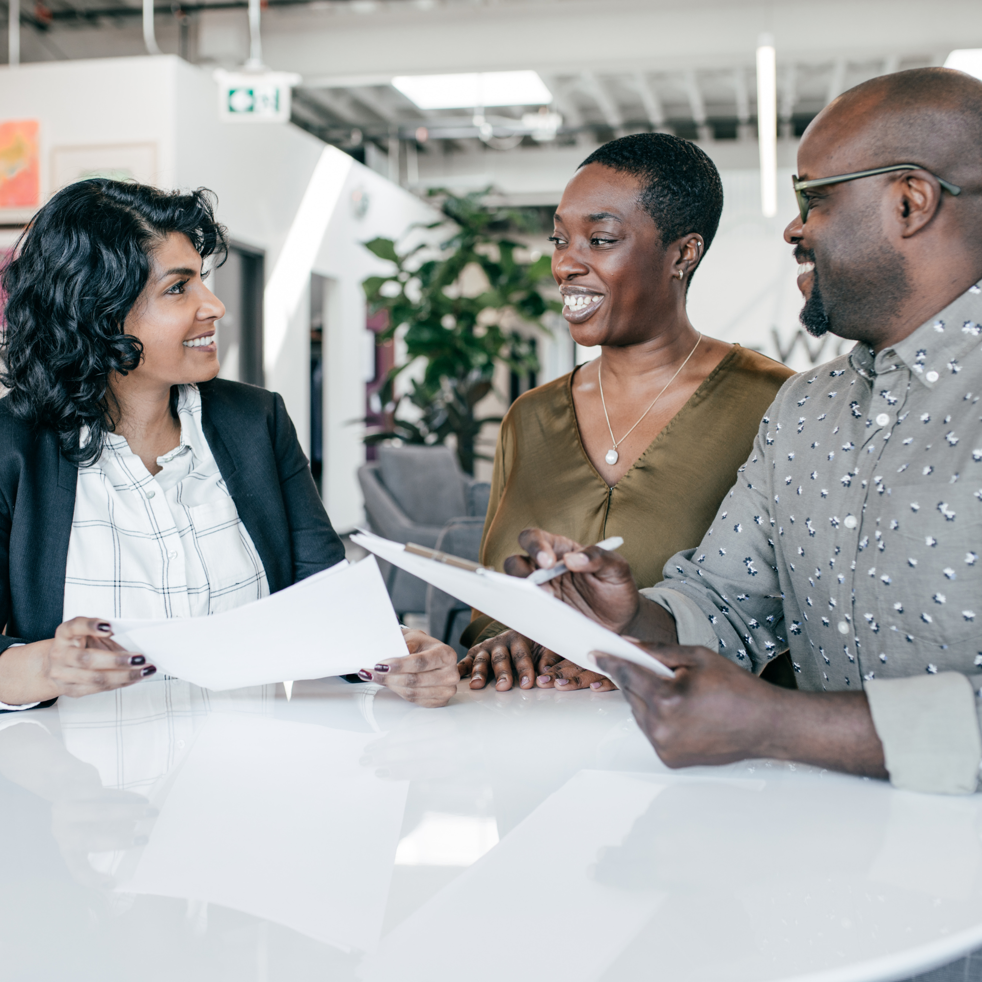 Three diverse professionals smiling and having a conversation in a modern office, holding papers and a tablet.