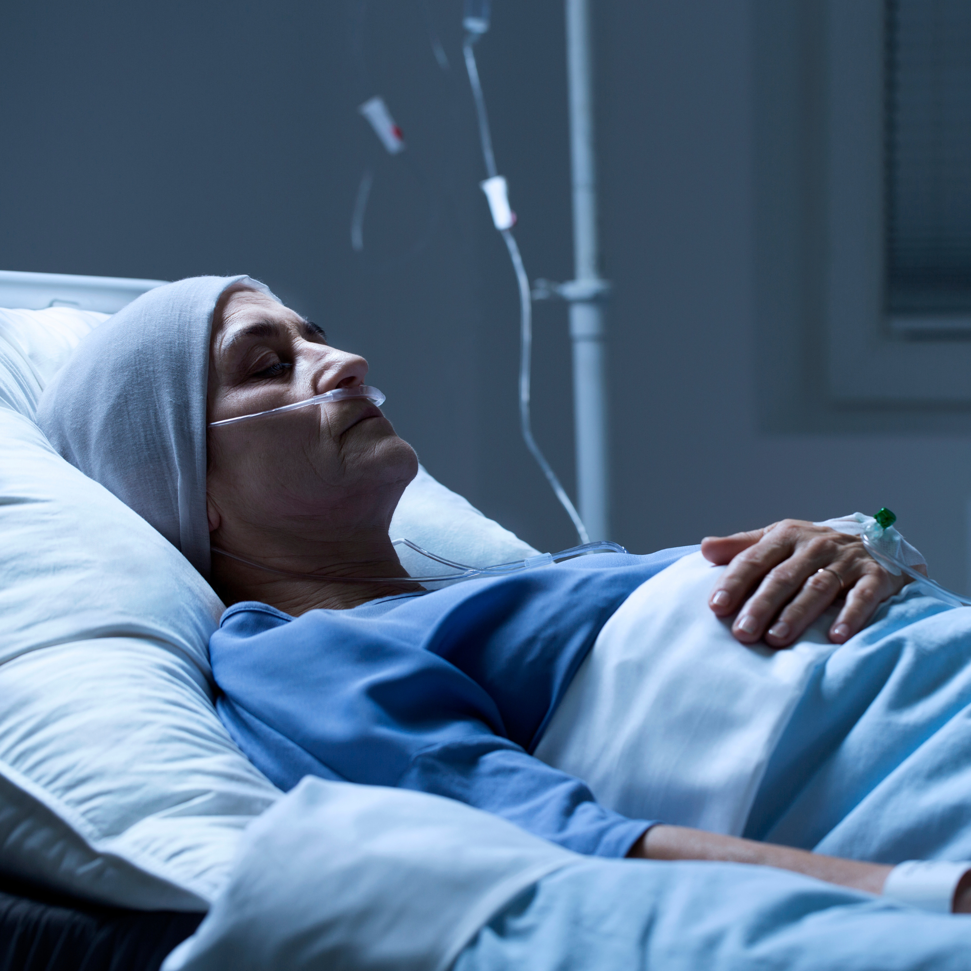 A patient lying in a hospital bed with a bandaged head, wearing a nasal oxygen tube and an intravenous drip, in a hospital room.