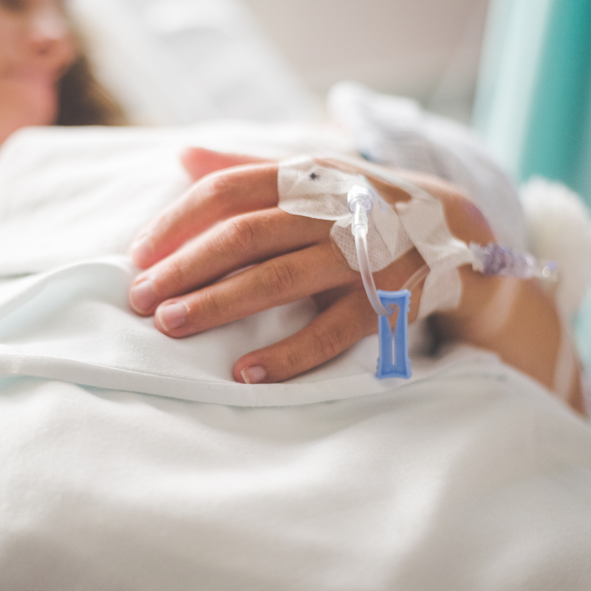 Close-up of a person’s hand holding a medical device with an IV line attached, a bandage around the finger, and a hospital or clinical setting in the background.