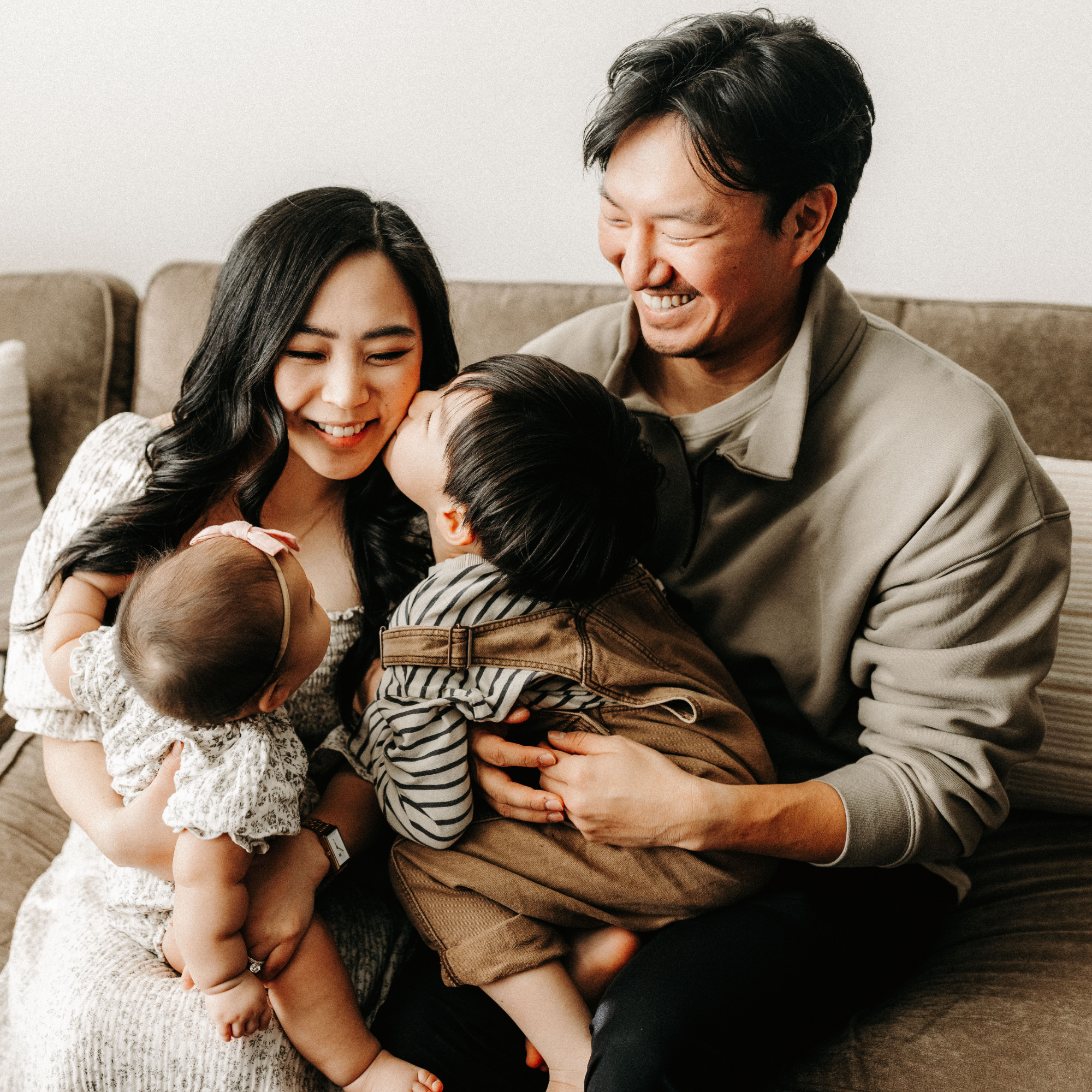 A happy family of four, including a father, mother, and two children, sharing a moment of affection and joy while sitting on a beige couch in a cozy living room.