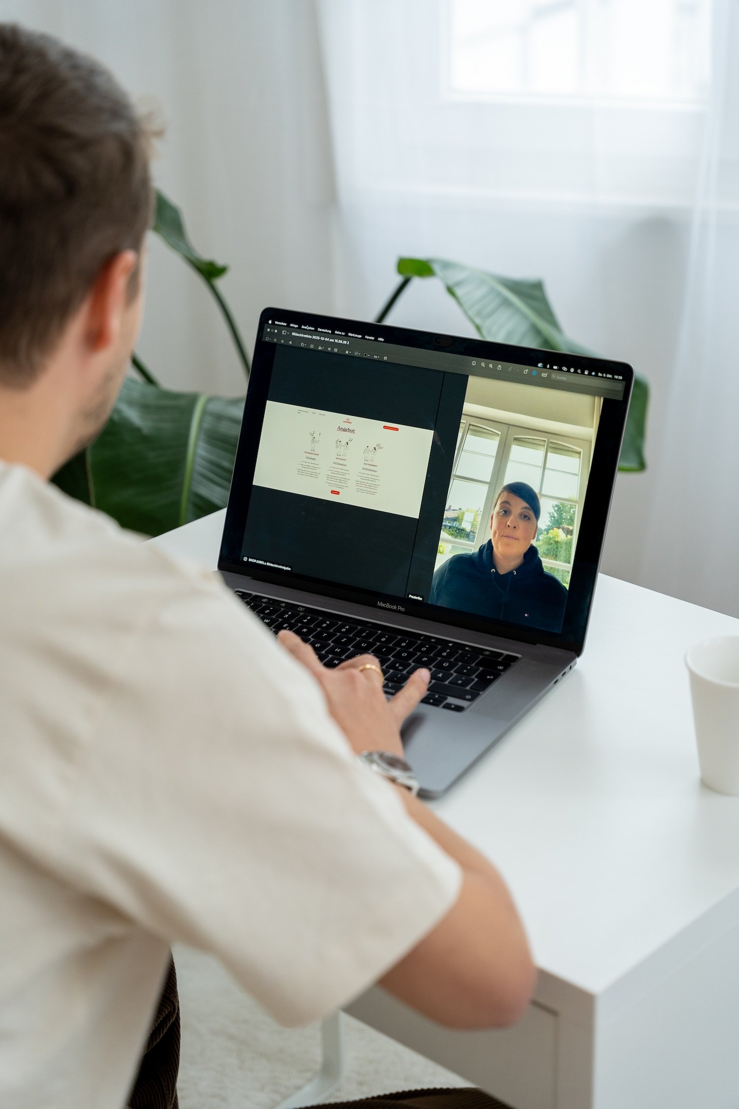 Eine Person arbeitet an einem Laptop, während sie an einem Videoanruf teilnimmt. Im Hintergrund sind Fenster und große grüne Pflanzen sichtbar.