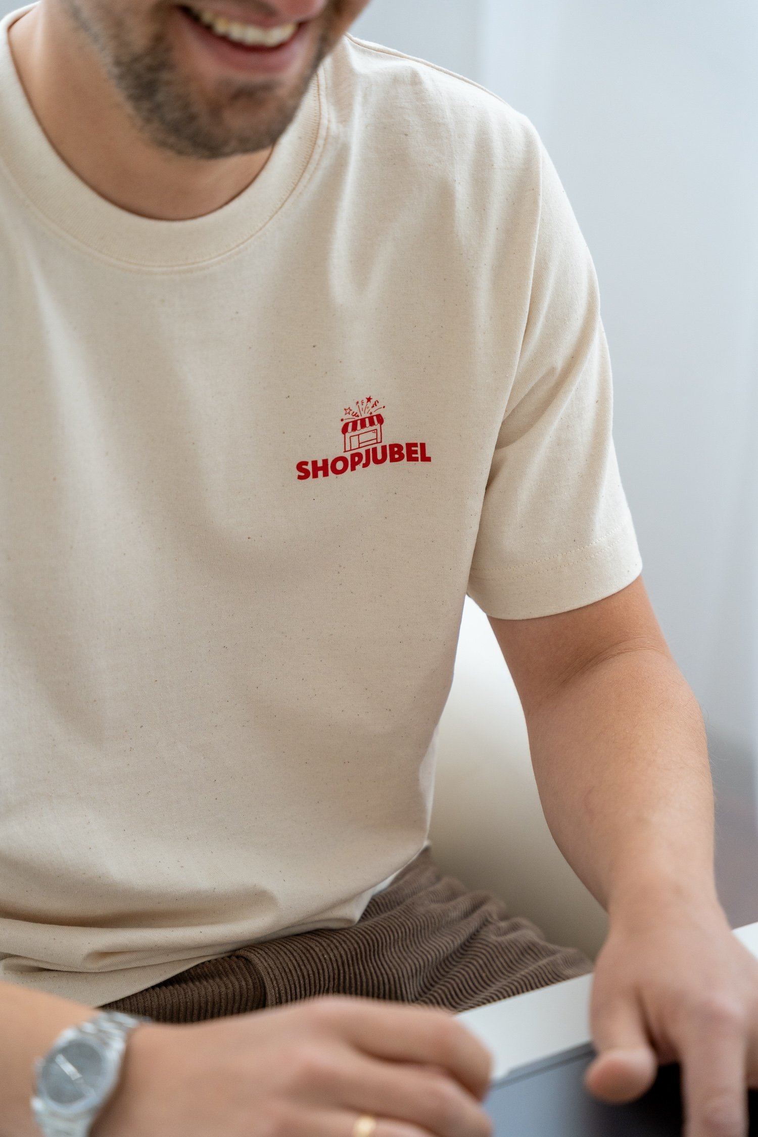 Nahaufnahme eines Mannes, der ein cremefarbenes T-Shirt trägt mit einem roten Aufdruck, der sagt "SHOPJUBEL" und ein Symbol eines Geschenkpakets mit Konfetti. Der Mann lächelt und hat einen Schmuck-Armband und eine goldene Ring an der Hand.