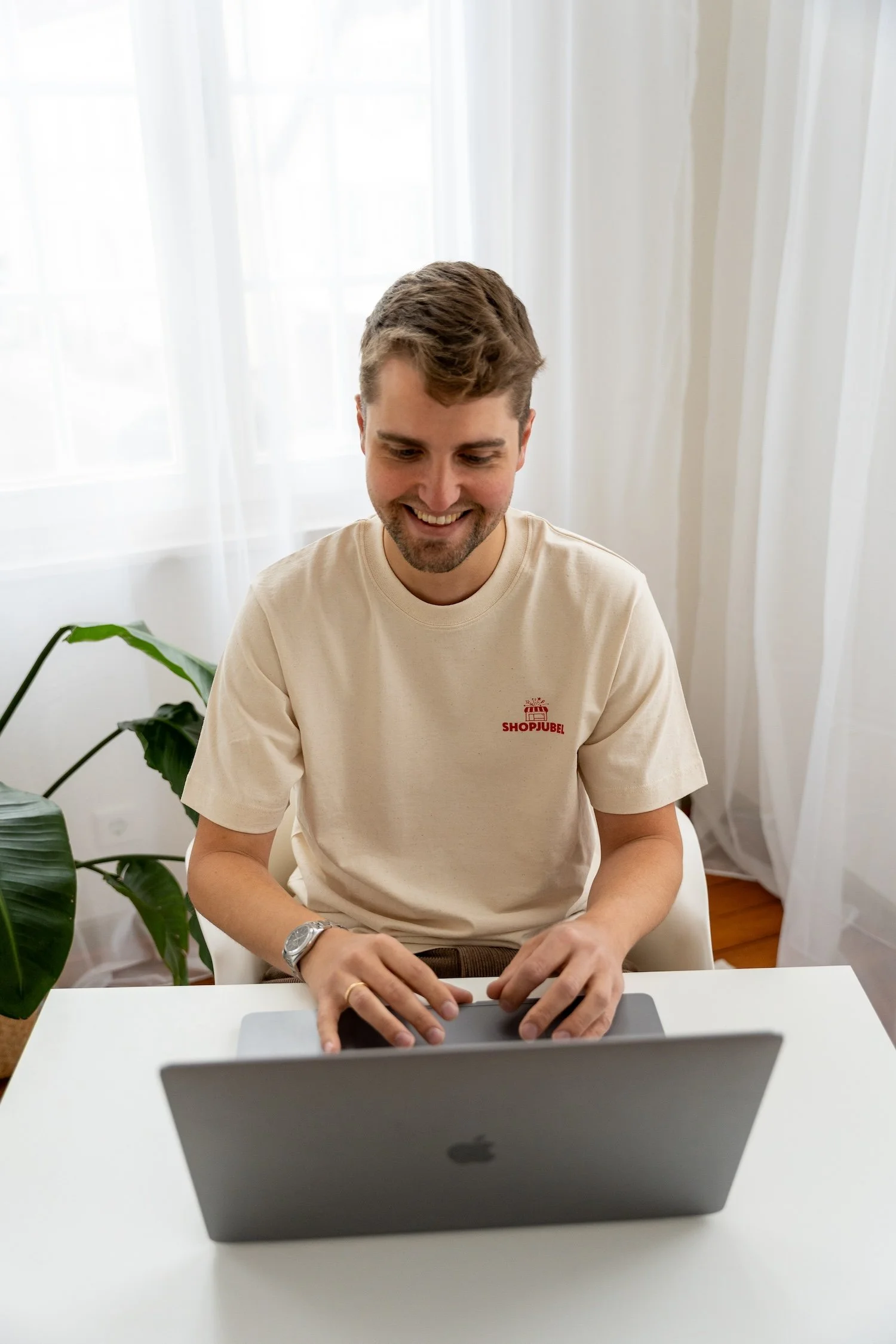 Junge Mann sitzt am Schreibtisch und benutzt einen Laptop in einem hellen Raum mit Vorhängen.