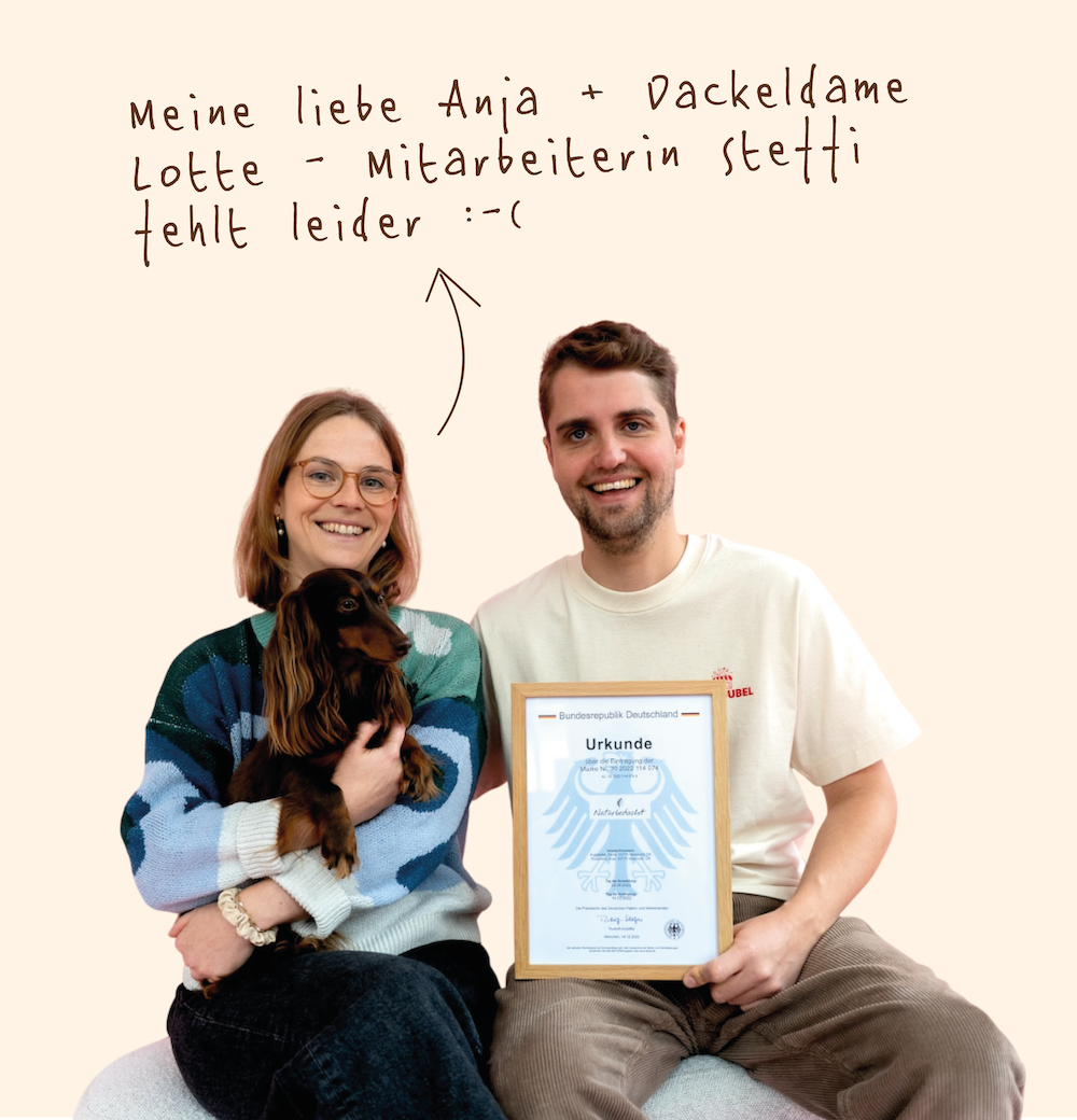 Ein glückliches Paar mit einem Hund, das ein Zertifikat in den Händen hält. Über dem Paar ist handgeschrieben in Deutsch: 'Meine liebe Anja + Dackeldame Lotte - Mitarbeiterin Stettl. tut leider :-('.