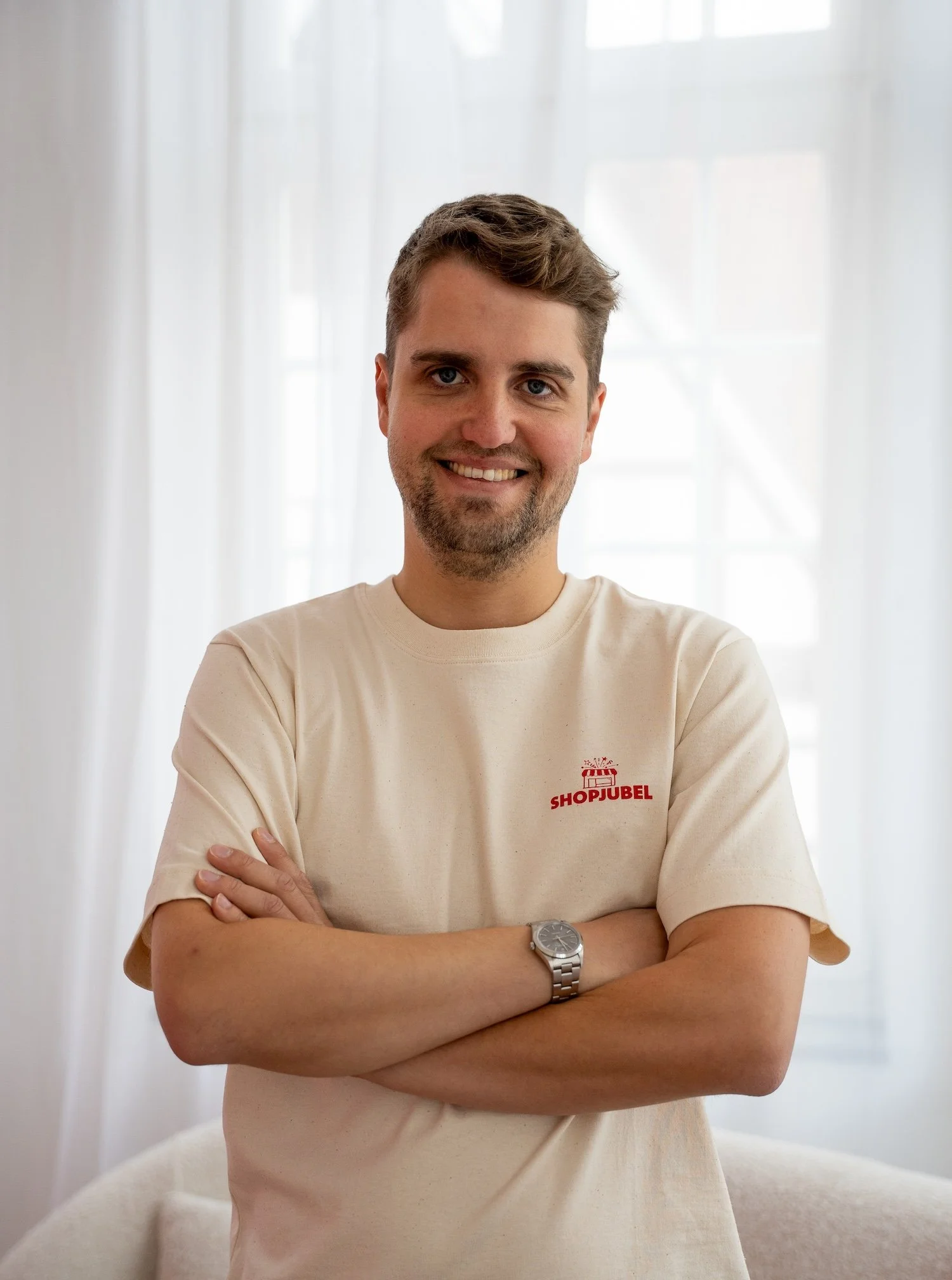 Ein Mann steht vor einem weißen Vorhang, trägt ein beige T-Shirt mit einem roten Logo und hat die Arme verschränkt. Er lächelt freundlich.