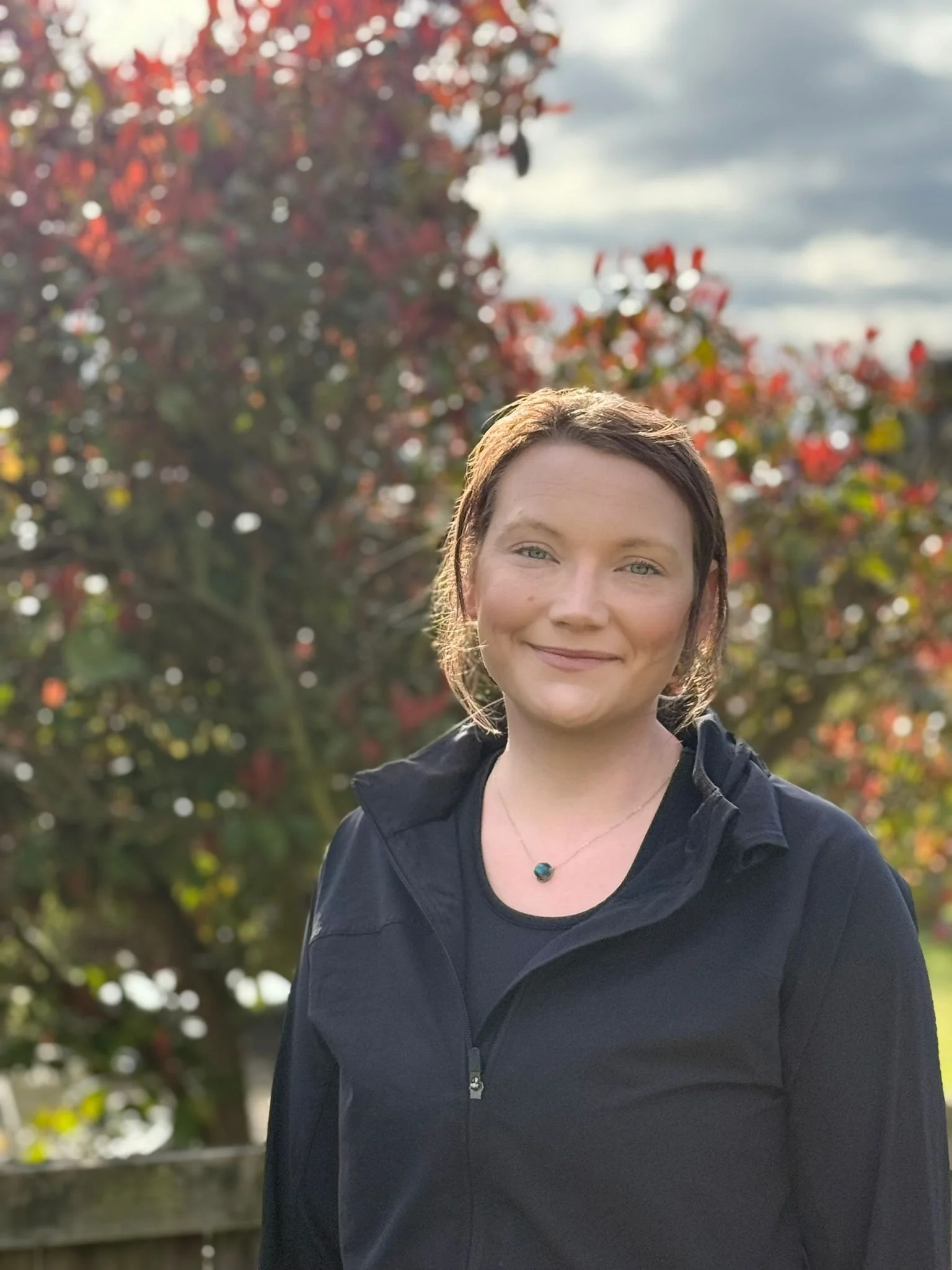 A woman with short brown hair and light skin smiling outdoors, wearing a black jacket and a necklace with a round pendant, standing in front of a tree with red leaves and a cloudy sky.