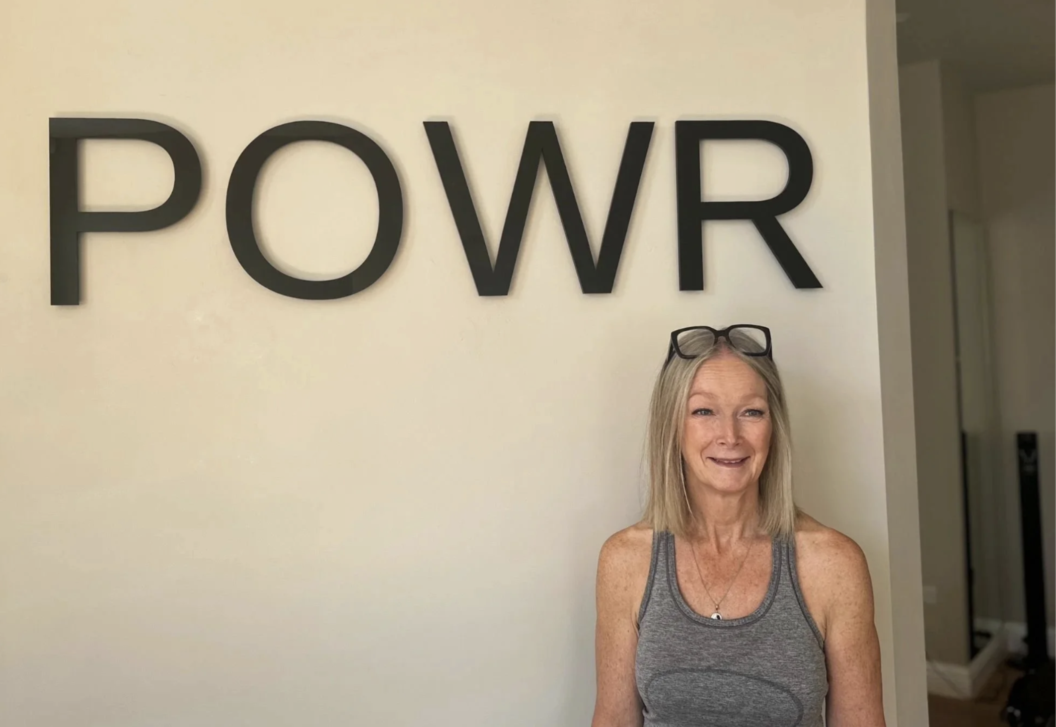 A woman with blonde hair wearing a gray sleeveless top and reading glasses on her head, standing in front of a wall with large black letters spelling 'POWER'.