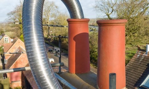 Metal chimney liner and two large red chimney pipes on a rooftop, with trees and rooftops in the background.