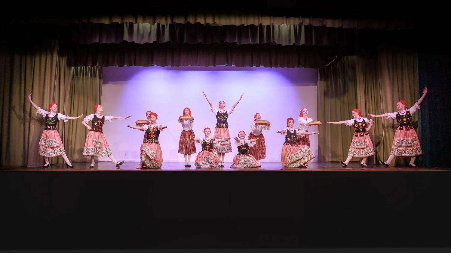 A group of dancers dressed in traditional folk costumes performing on stage, with some holding cake trays and others striking dance poses.