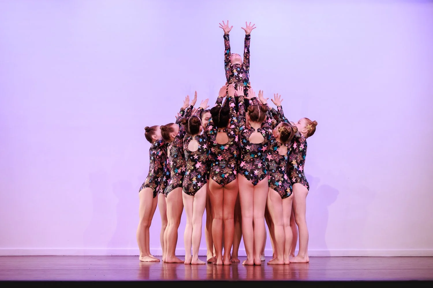 A group of dancers in colourful star-patterned costumes form a pyramid on stage, with one girl at the top reaching upward.