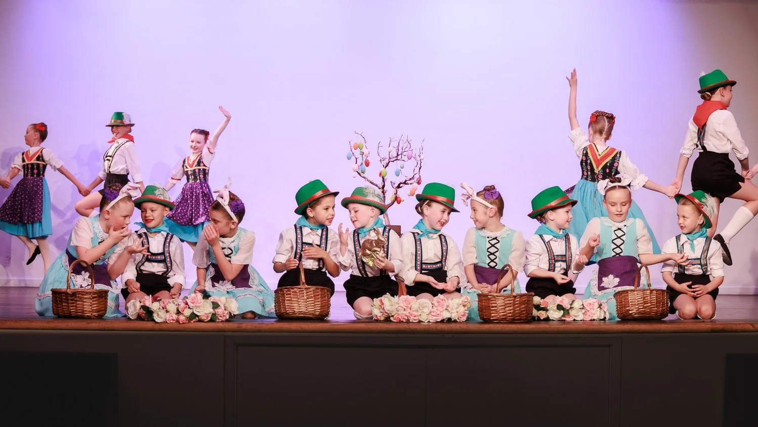 Children in traditional European costumes performing a dance on stage with a white backdrop, some sitting with baskets, others in pairs holding hands, and a decorative tree with hanging ornaments in the center.