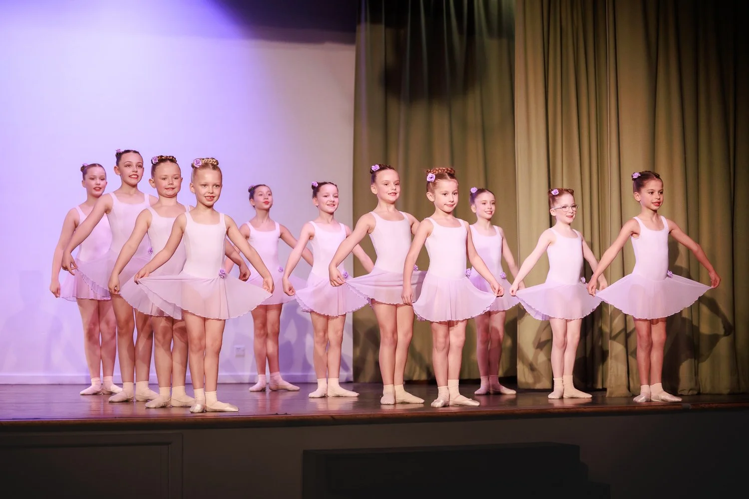 Group of ballet dancers in white tutus performing on stage with green curtains behind them.