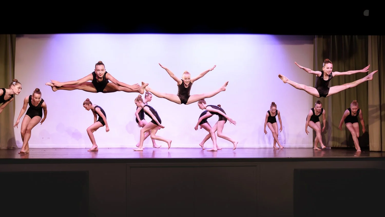 Group of dancers in black performing a dance routine on a stage, with some leaping and others preparing to move.
