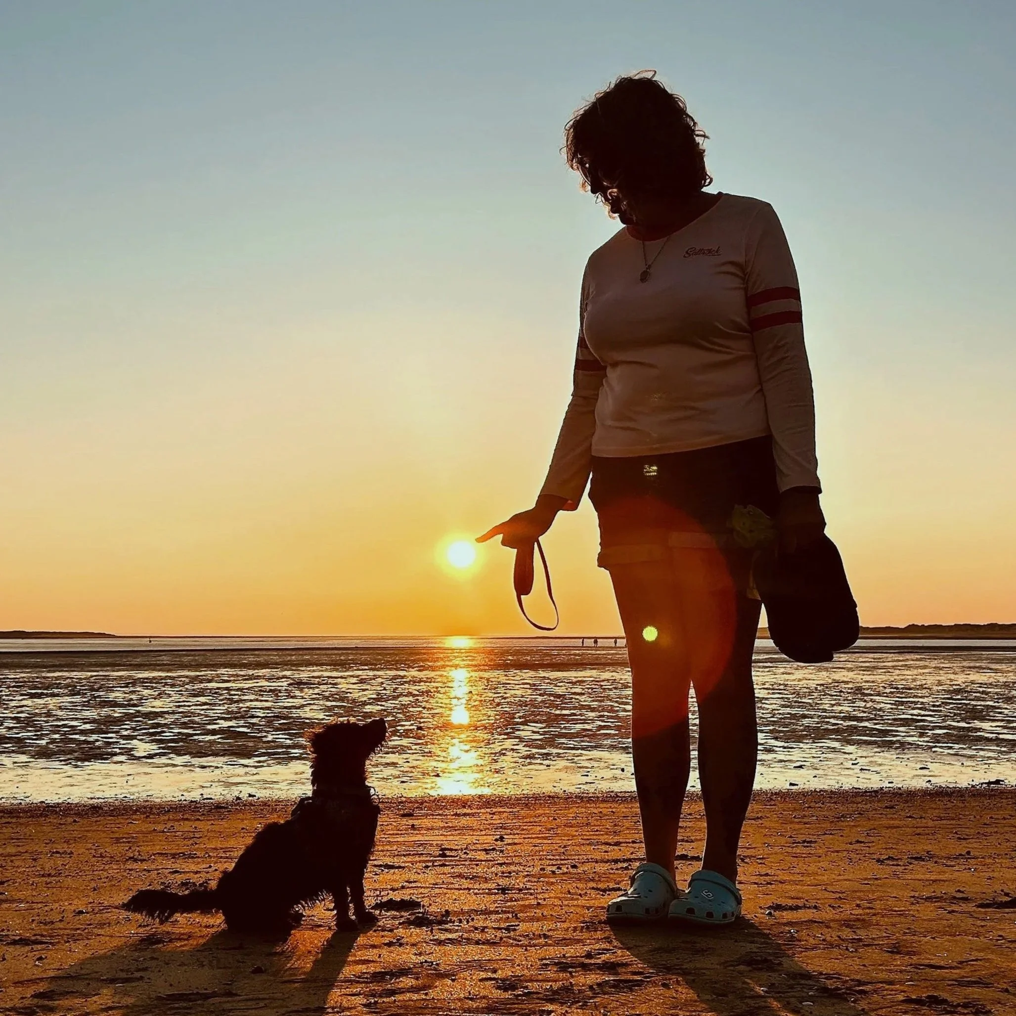 A woman with curly hair standing on the beach at sunset, holding a leash attached to a small dog sitting on the sand, with the ocean and sunset in the background.