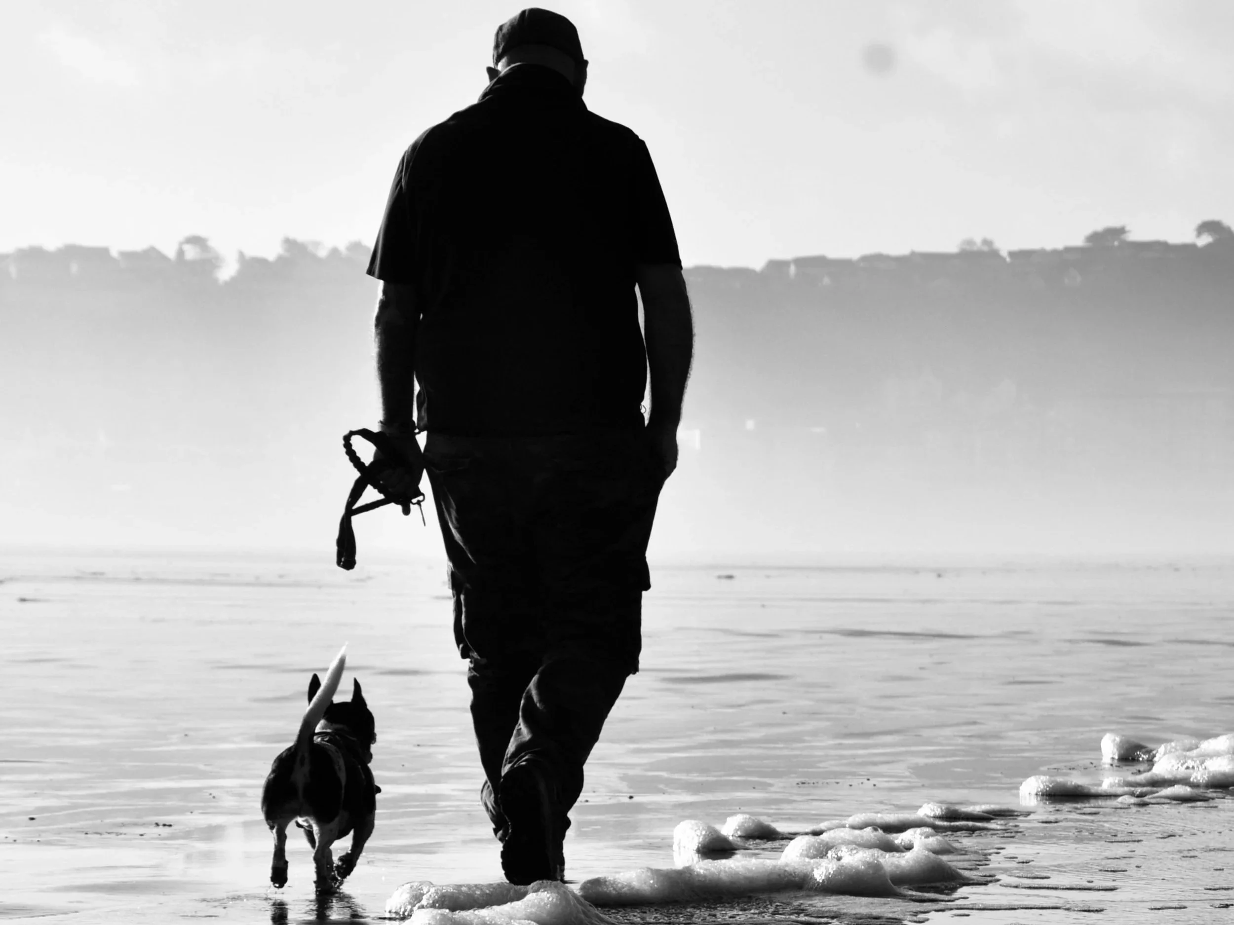 Silhouette of a person walking a dog along the shoreline, with waves and foam on the sand near the water in black and white.
