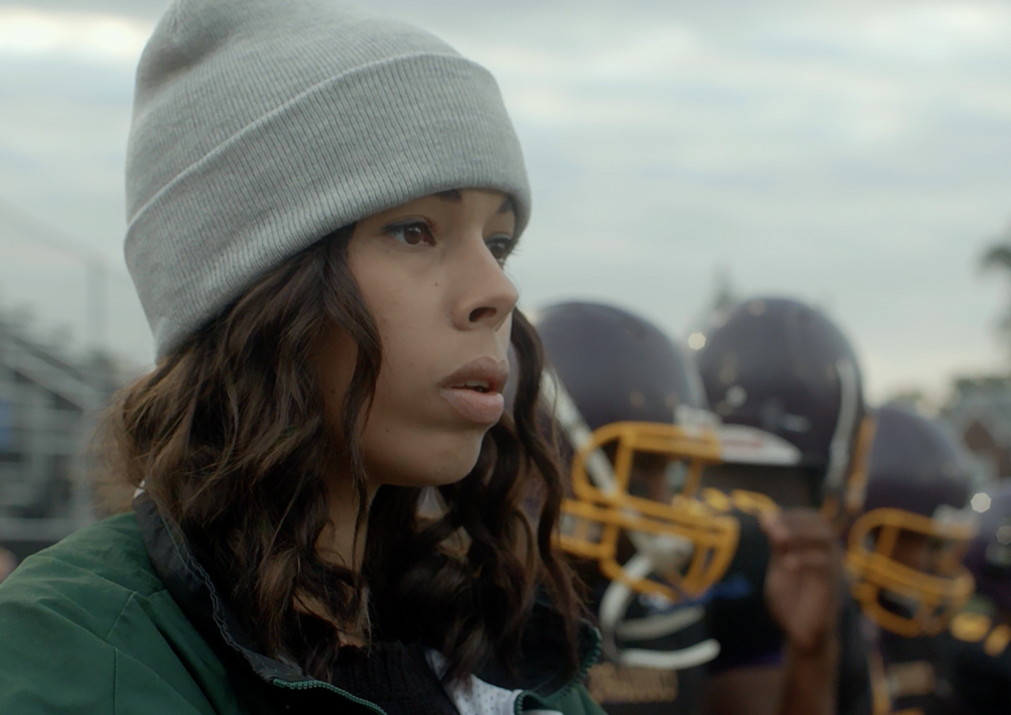 Close-up of a young woman wearing a gray beanie and green jacket, standing outdoors with a football team in the background.