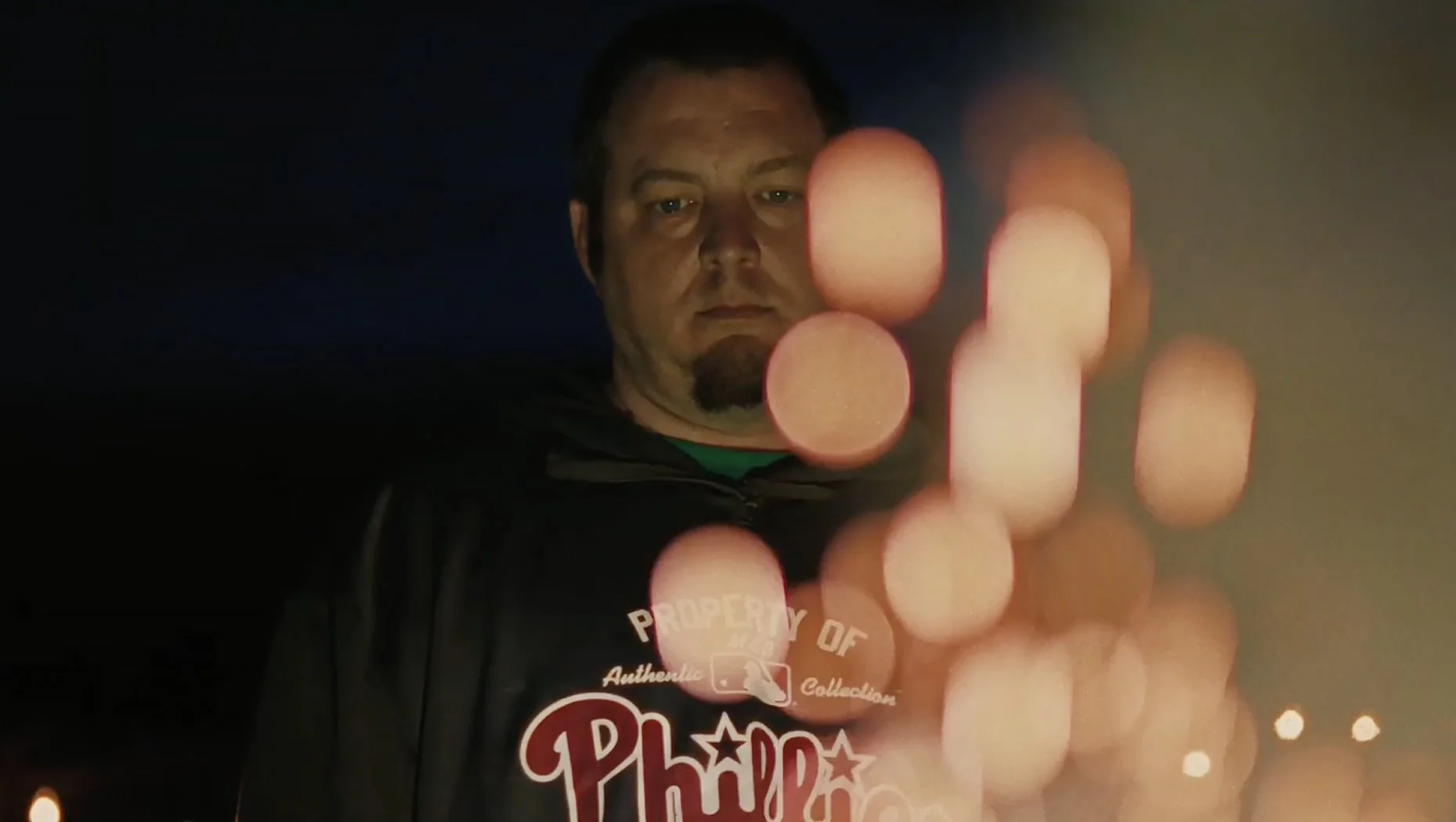 A man in a black hoodie with a logo, looking at the camera, with a background of blurred lights, and his finger appears to be touching a glass surface or screen that reflects or displays blurred circular light patterns.